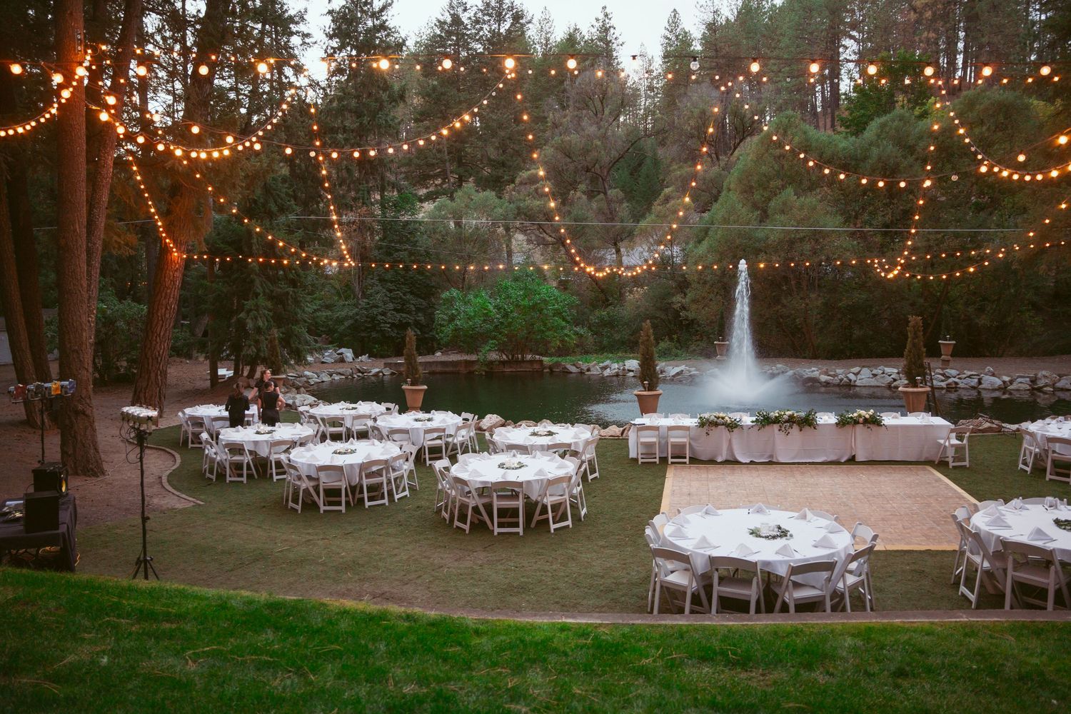 String lights illuminate an outdoor wedding reception setup with round tables and chairs in a wooded forest setting.