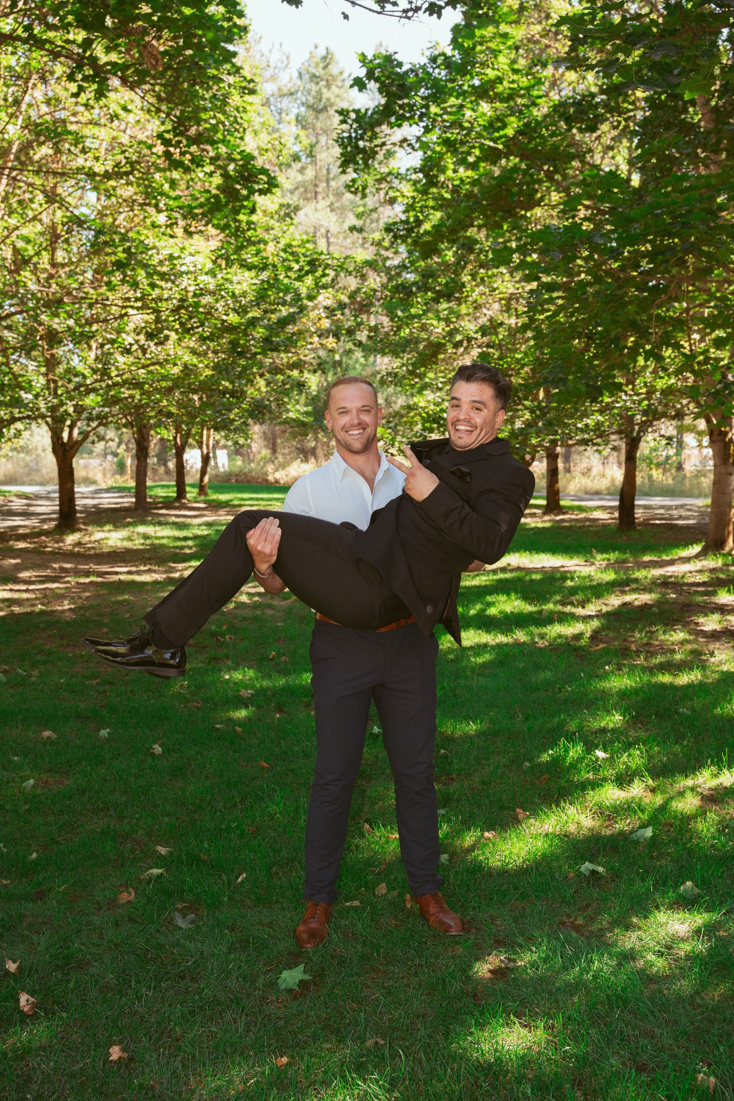 Two people in dark suits playfully pose in a grassy park with trees.