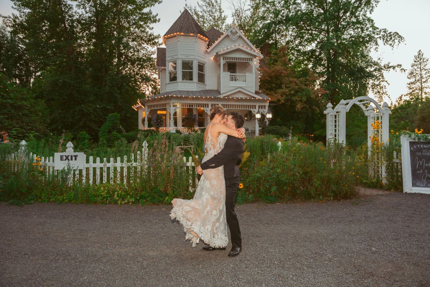 A romantic wedding couple embraces in front of a beautiful Victorian mansion with a white picket fence at dusk.