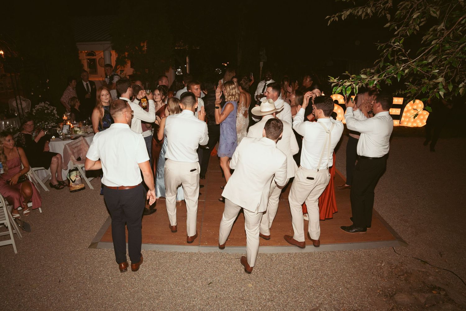 Group of wedding guests in white attire dancing together at outdoor reception.