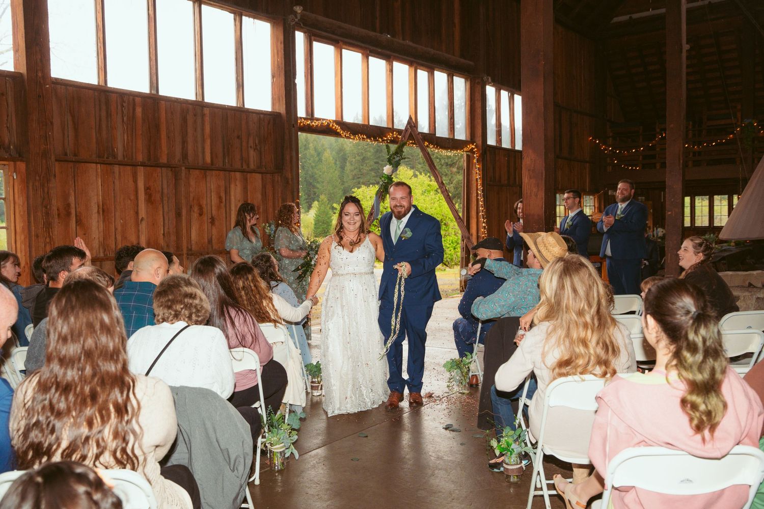 Full view of wedding ceremony in rustic barn with guests seated on both sides.