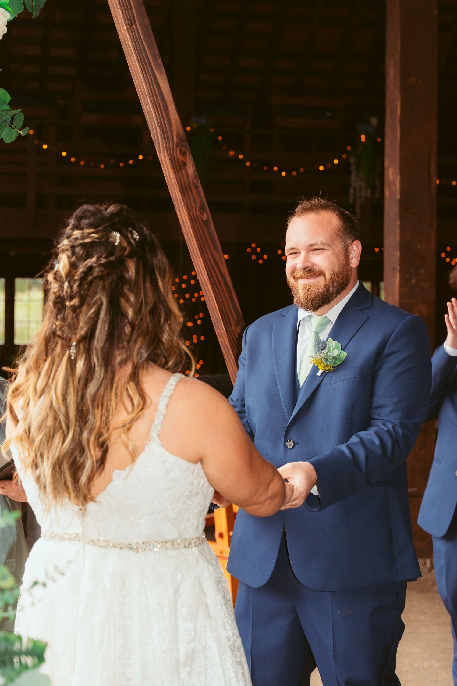 Couple exchanging vows during intimate barn wedding ceremony with string lights and greenery.