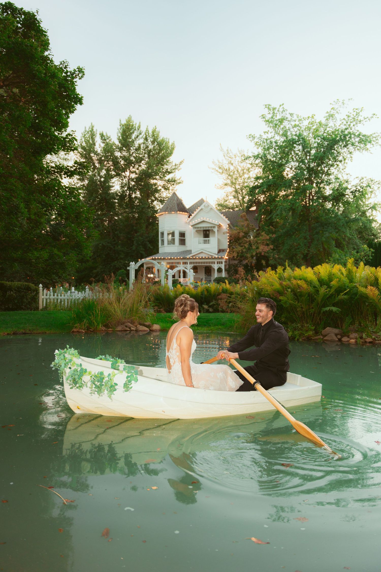 Couple rowing in a white wooden boat on a tranquil pond with a house in the background.