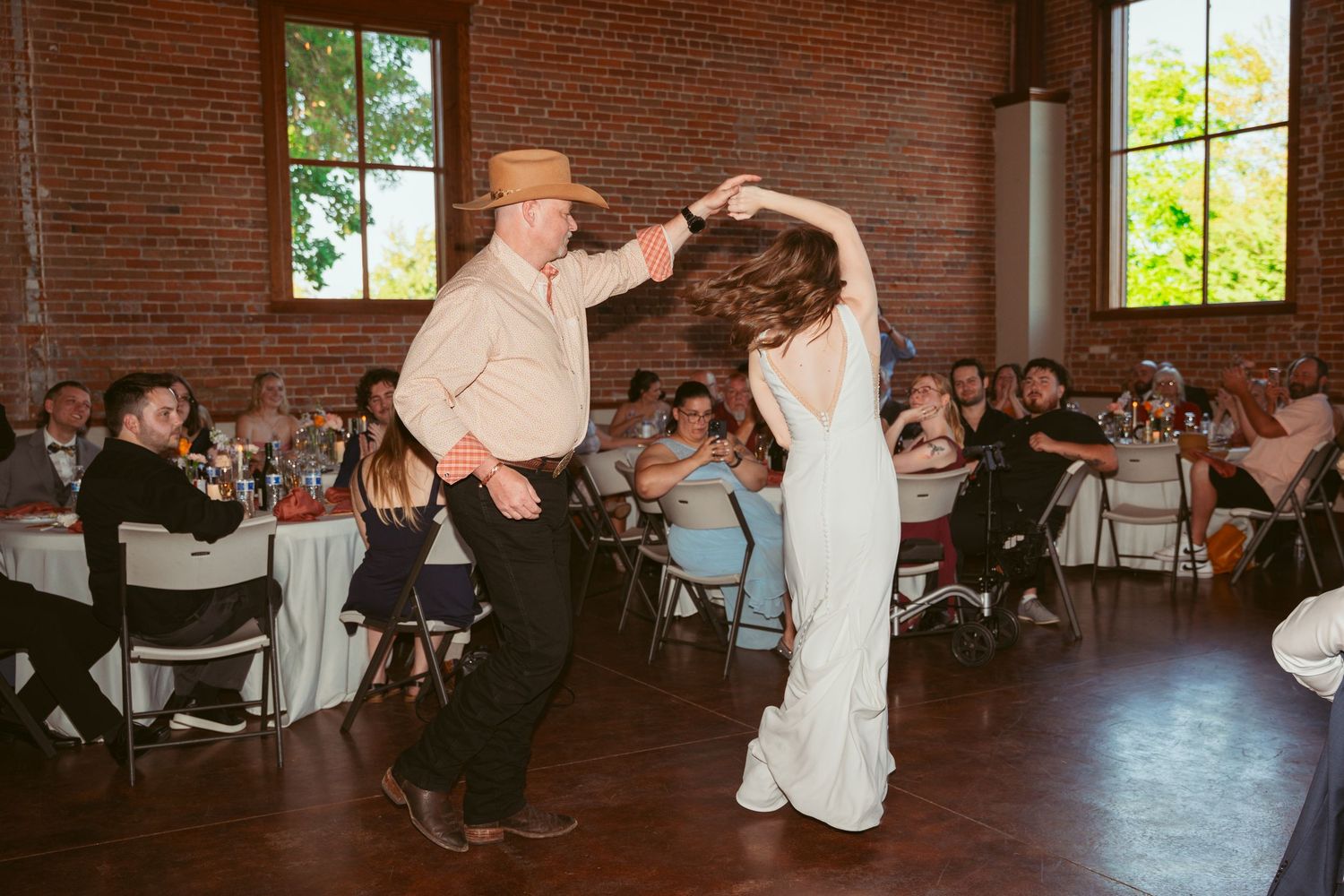 Dancers perform a lively twirl in front of wedding guests at an indoor brick venue with tall windows.