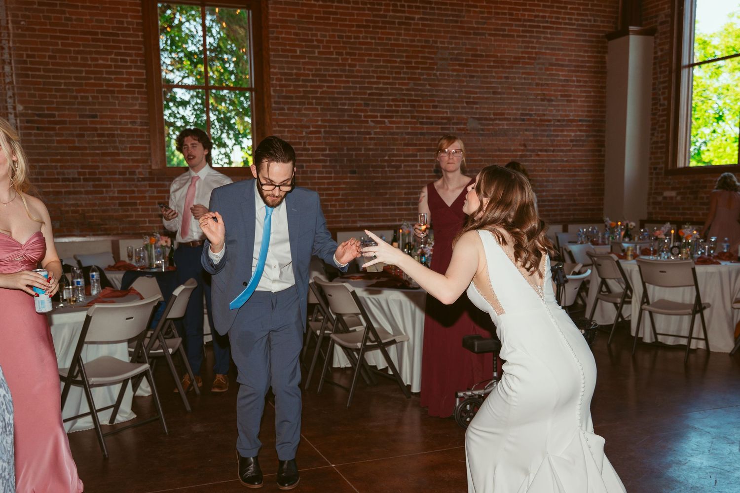 Wedding party dances enthusiastically in a spacious brick venue with folding chairs.