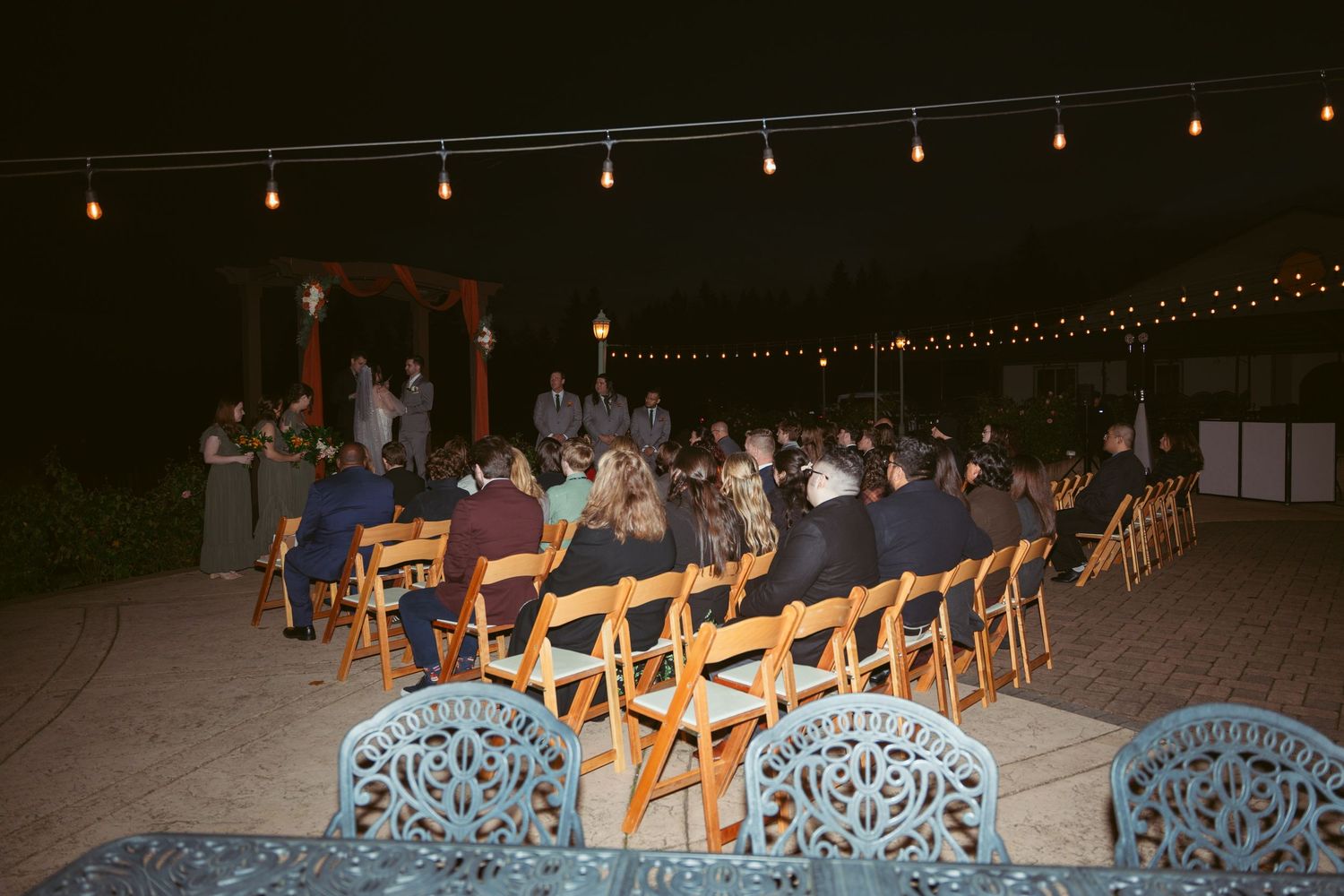 Wedding guests seated in rows of wooden chairs at an outdoor evening reception with string lights overhead.