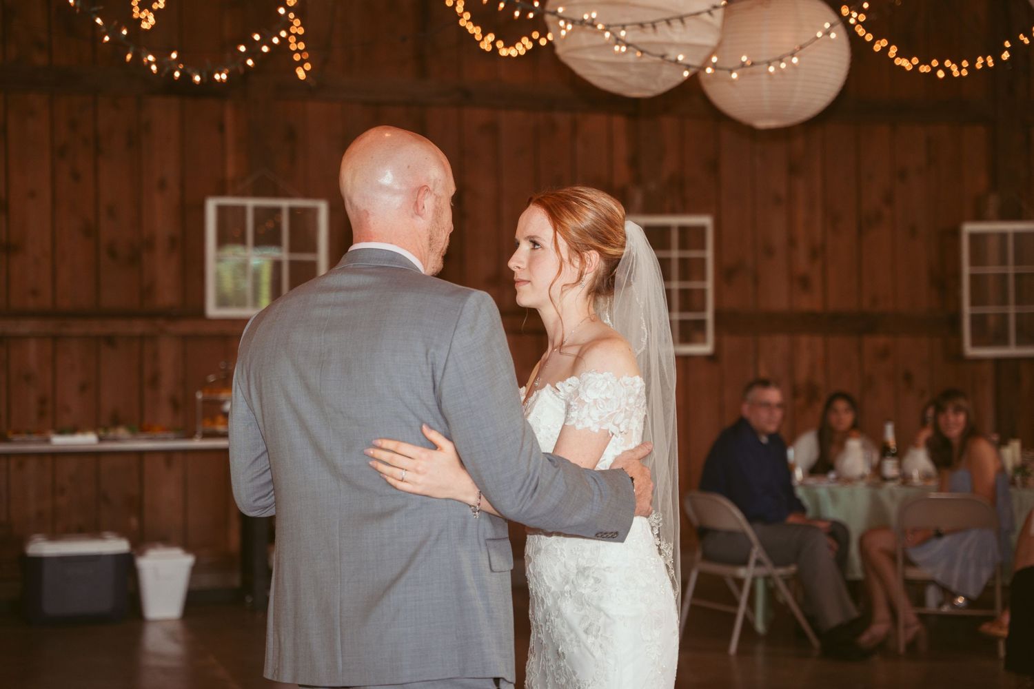 A couple shares their first dance in a rustic barn venue decorated with twinkling string lights and paper lanterns.