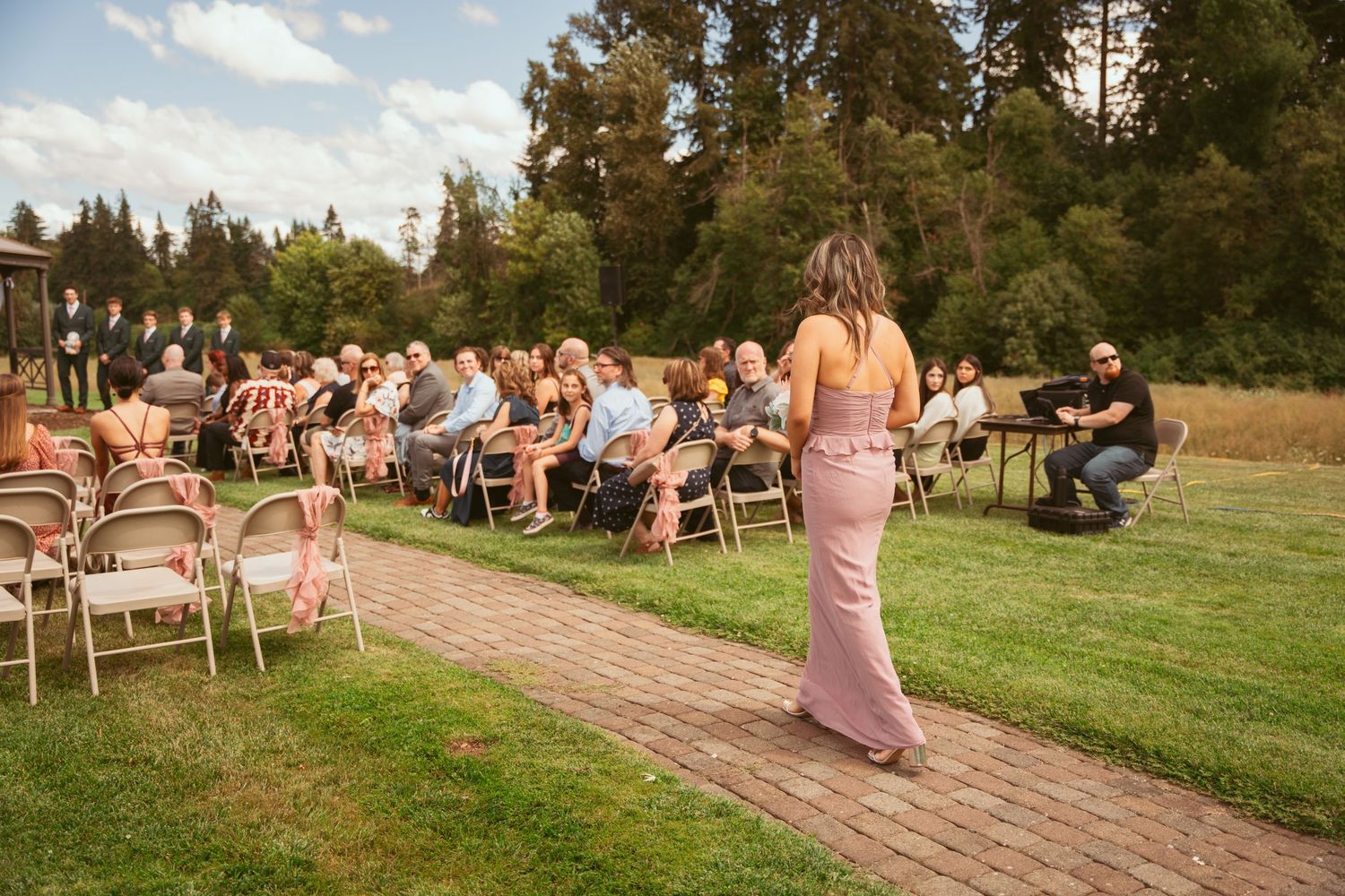Wedding guests seated on chairs watch as someone walks down a brick aisle outdoors.