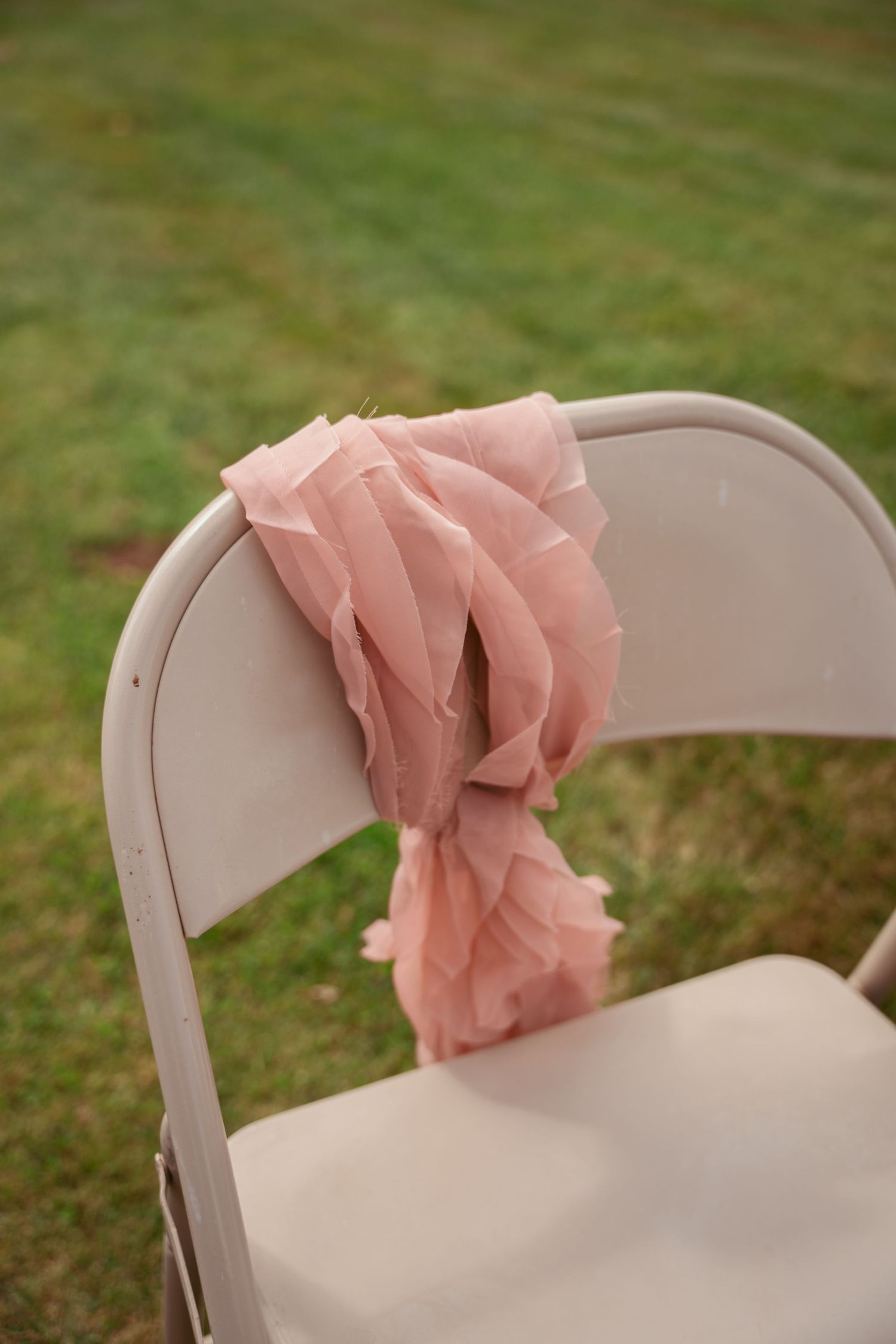 White folding chairs decorated with blush pink fabric bows set up for outdoor ceremony.
