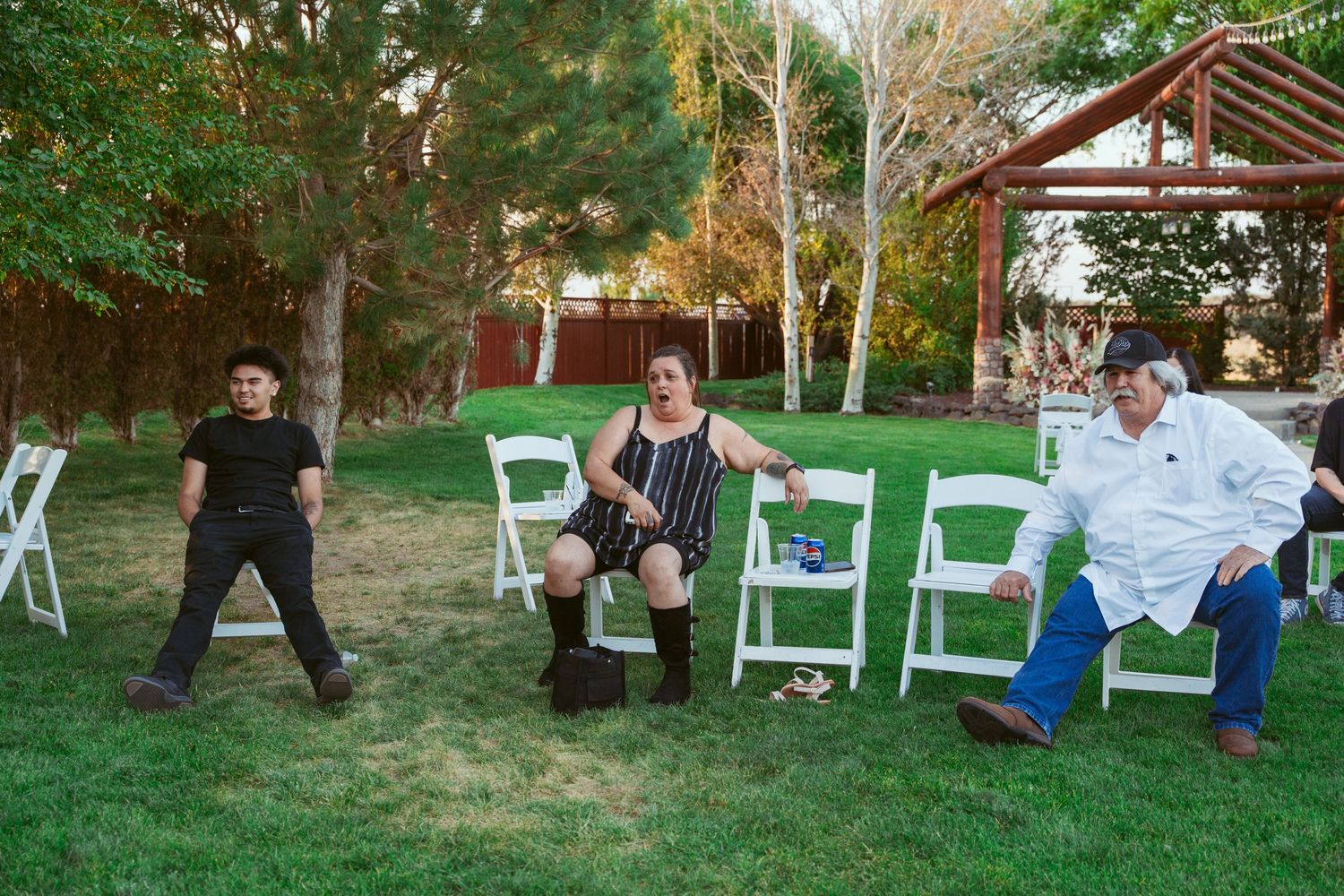 Wedding guests dance and celebrate outdoors on a lawn with white chairs in the background during daytime.