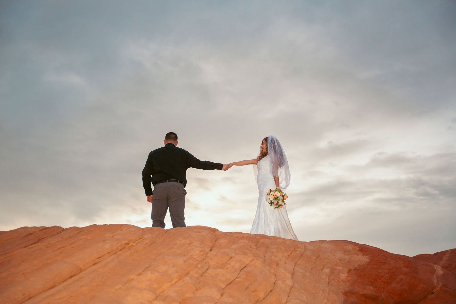 Silhouettes of newlyweds stand against a cloudy sky on red desert rocks.