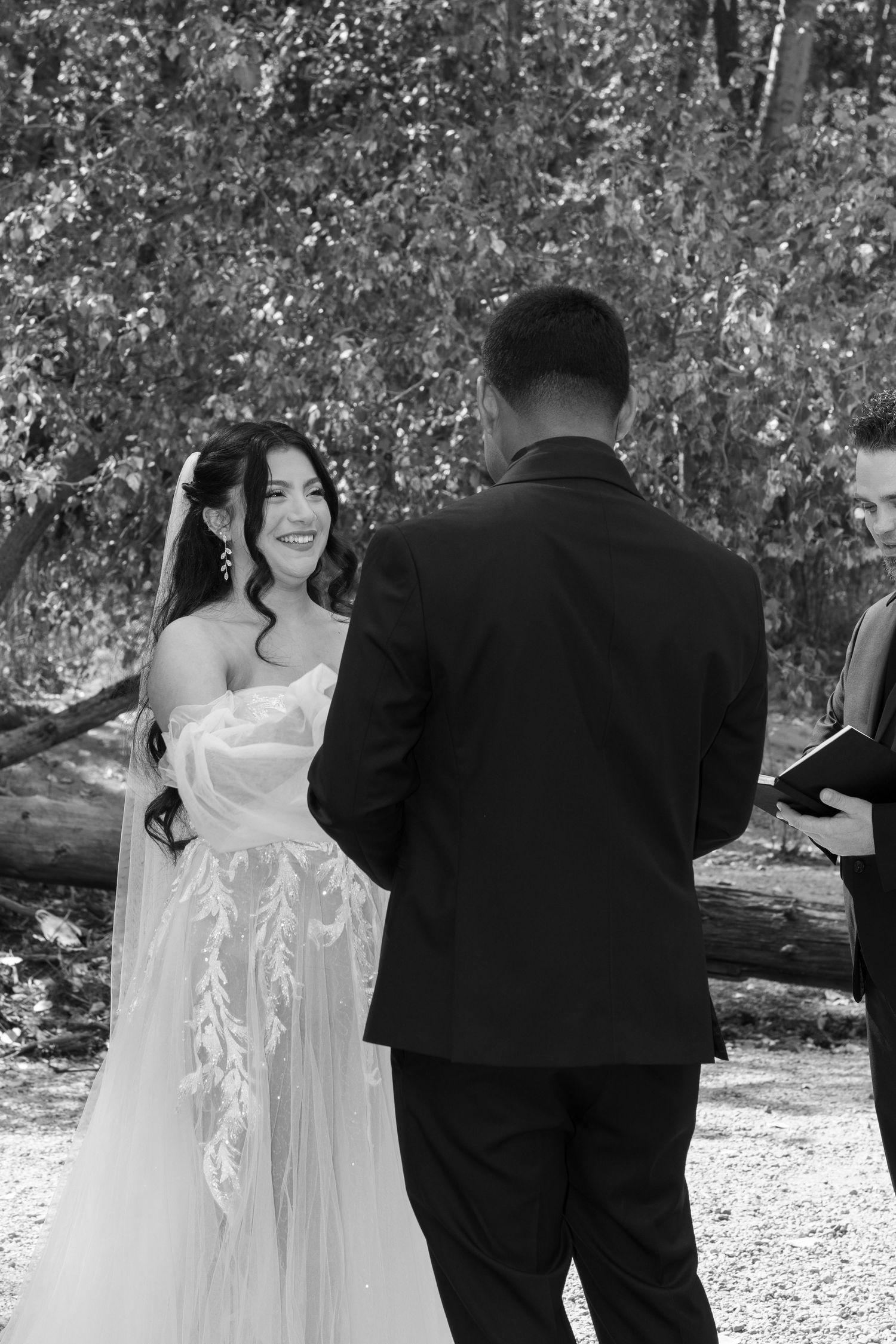 A bride and groom exchange vows during their black and white wedding ceremony surrounded by trees.