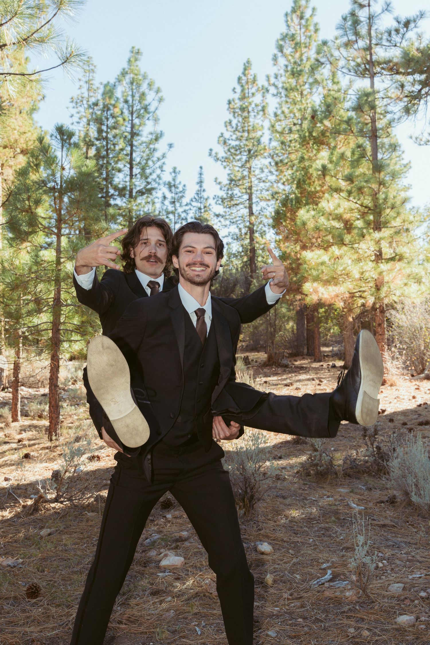 Two people in suits playfully pose in a forest clearing.