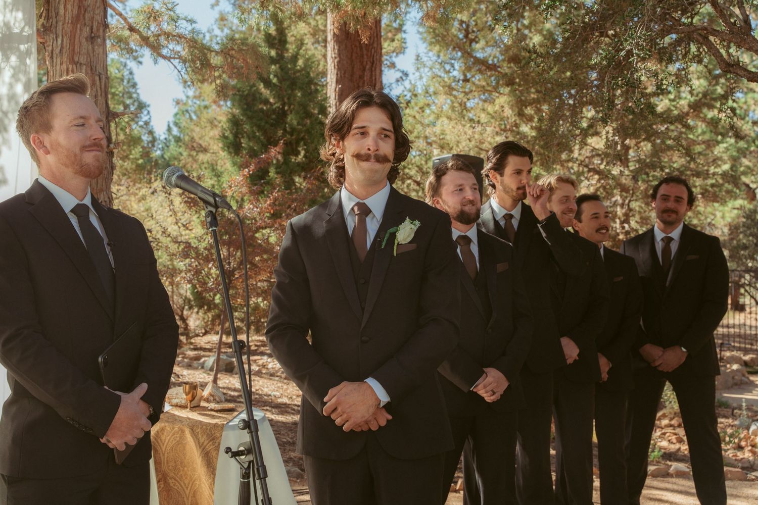 Groomsmen in dark suits standing in formation at an outdoor wedding ceremony.