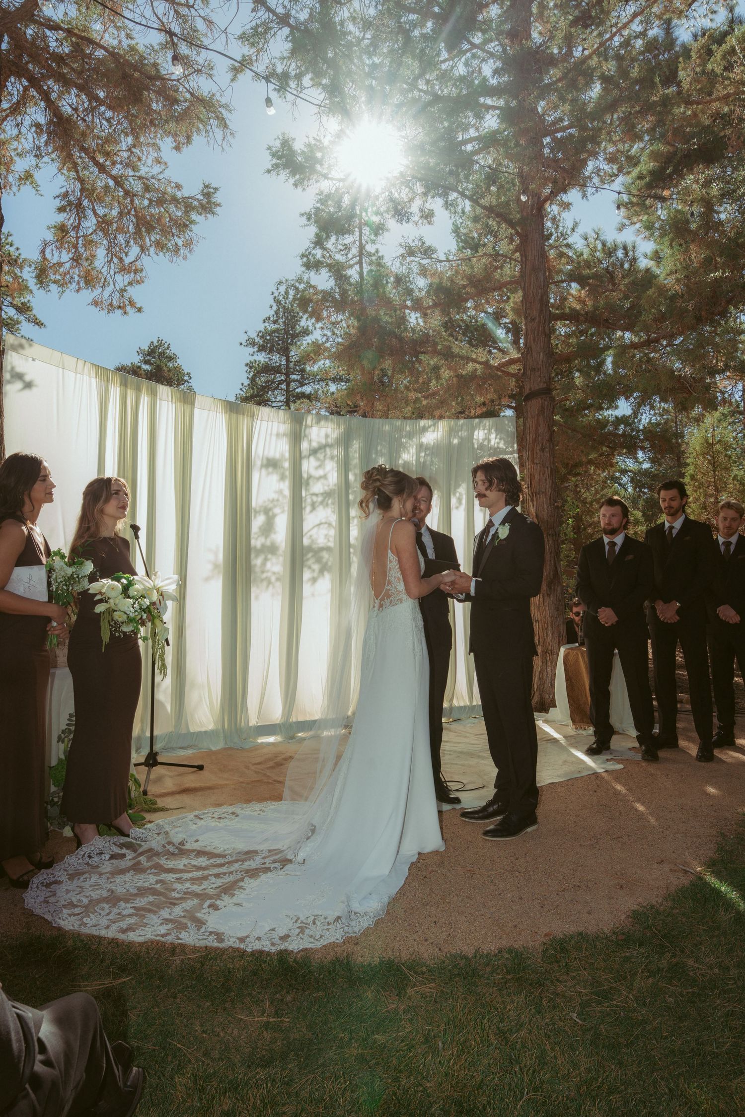 Wedding ceremony in a sunlit forest setting with white drapes and wedding party standing in formation.