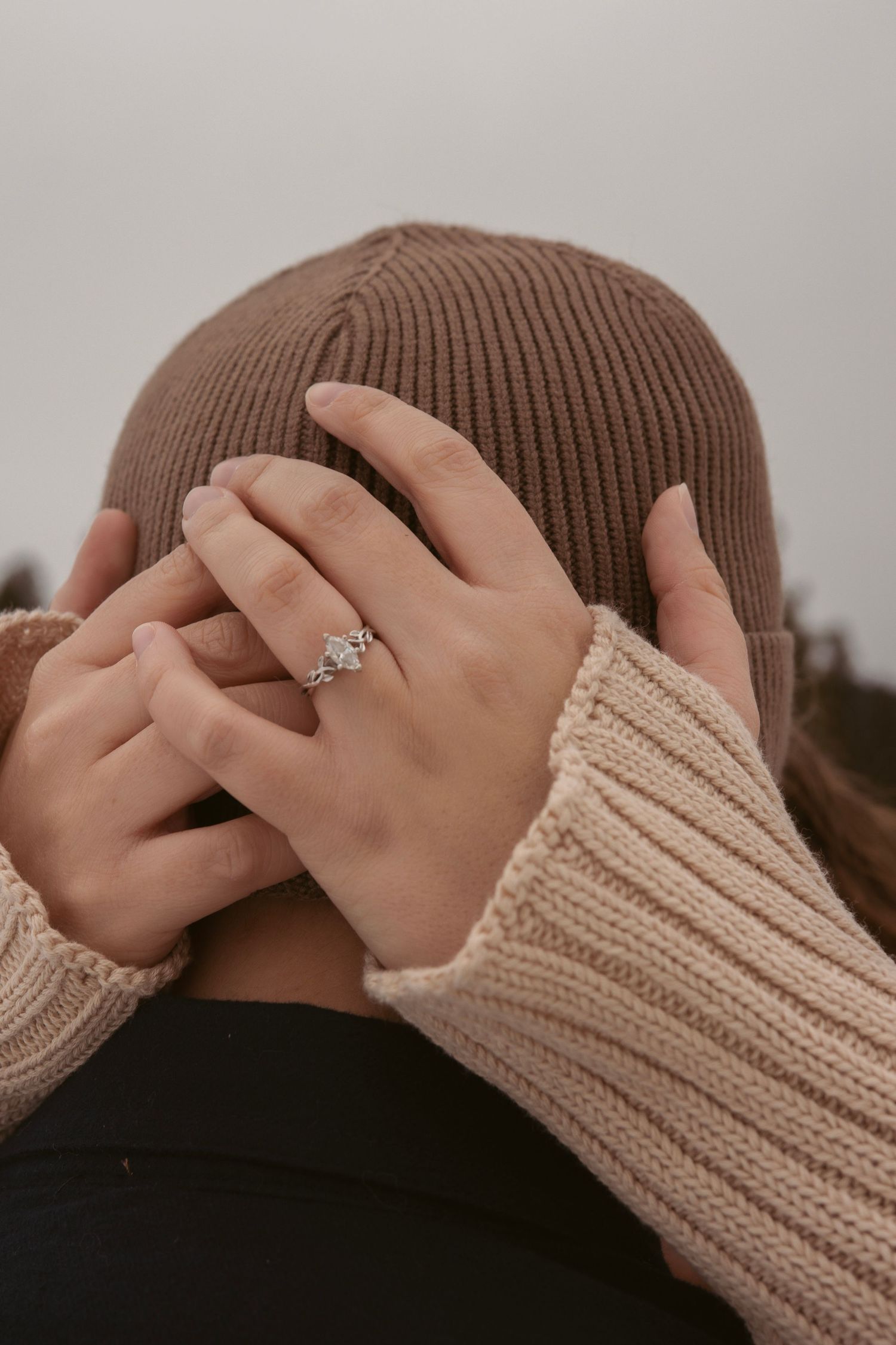Close-up of hands wearing knit sweater and brown beanie showing engagement ring.