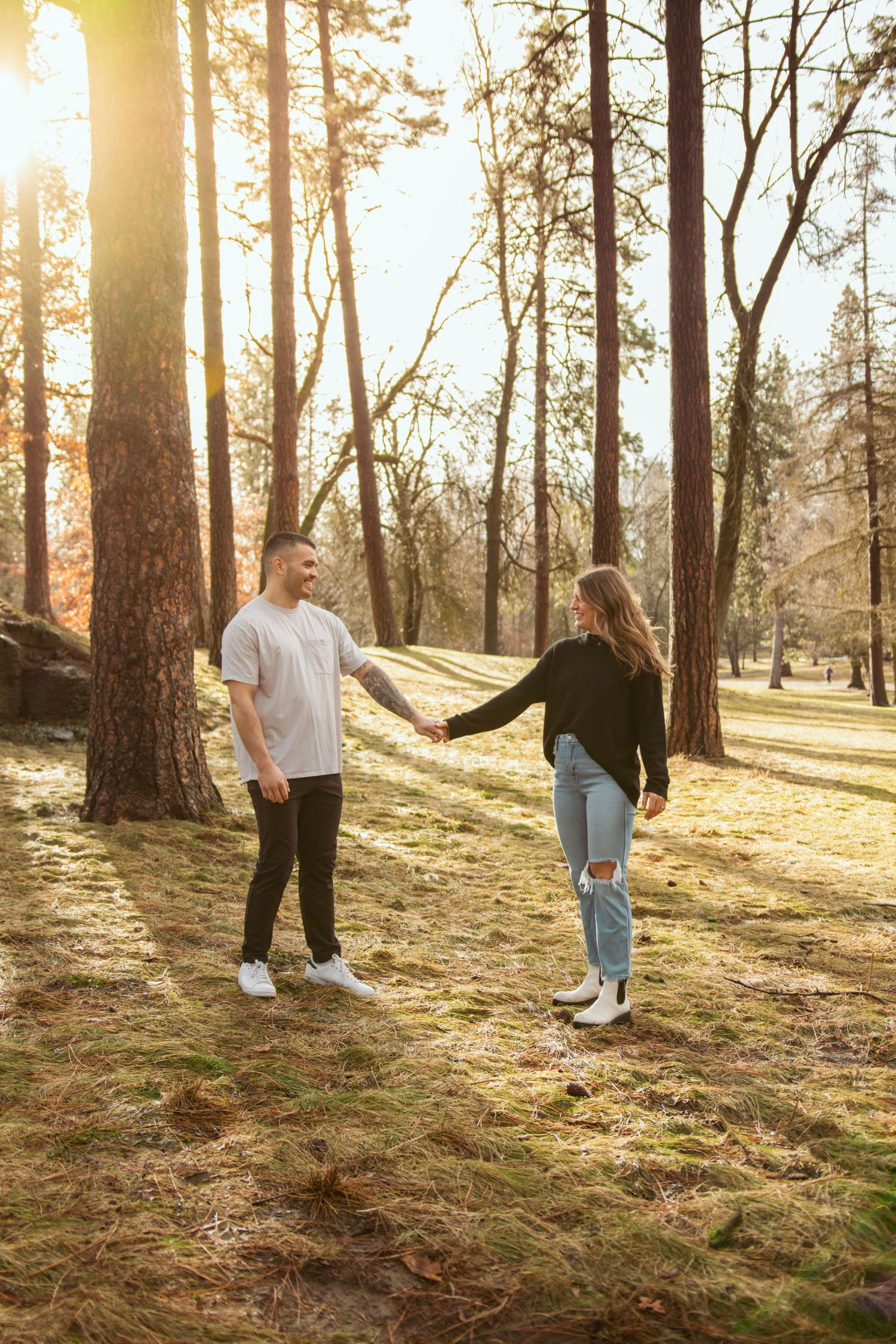 A couple walks playfully through a sunlit forest with tall pine trees.