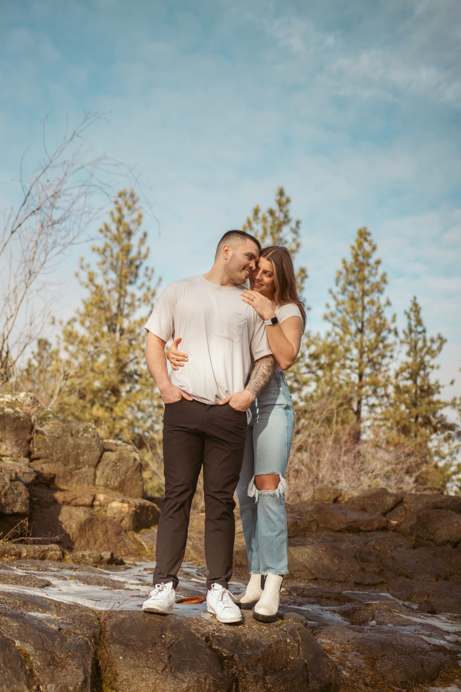 Couple standing together on rocky terrain with evergreen trees in the background.