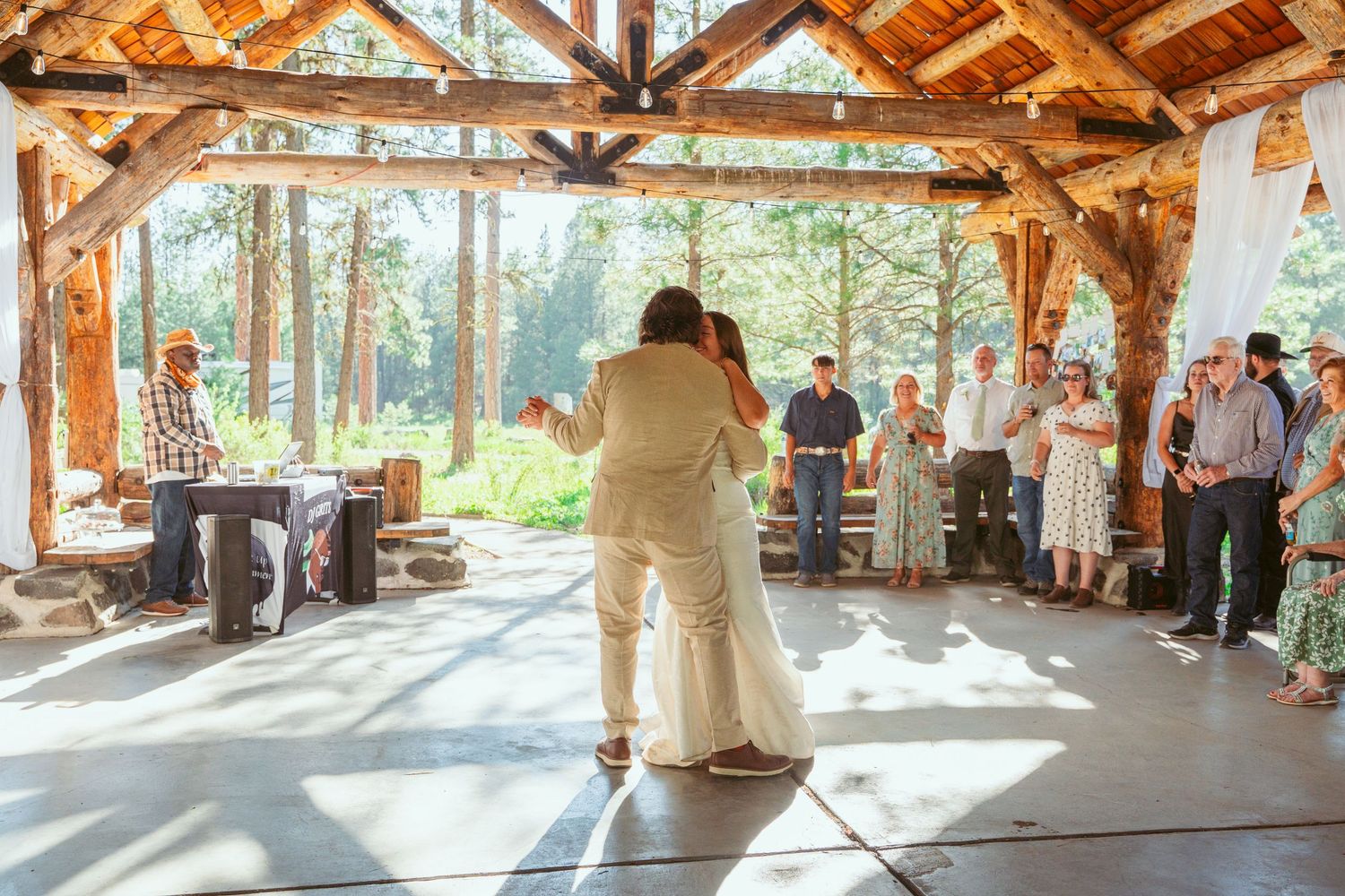 Couple dancing together in a sunlit wooden pavilion with guests watching from the sides.
