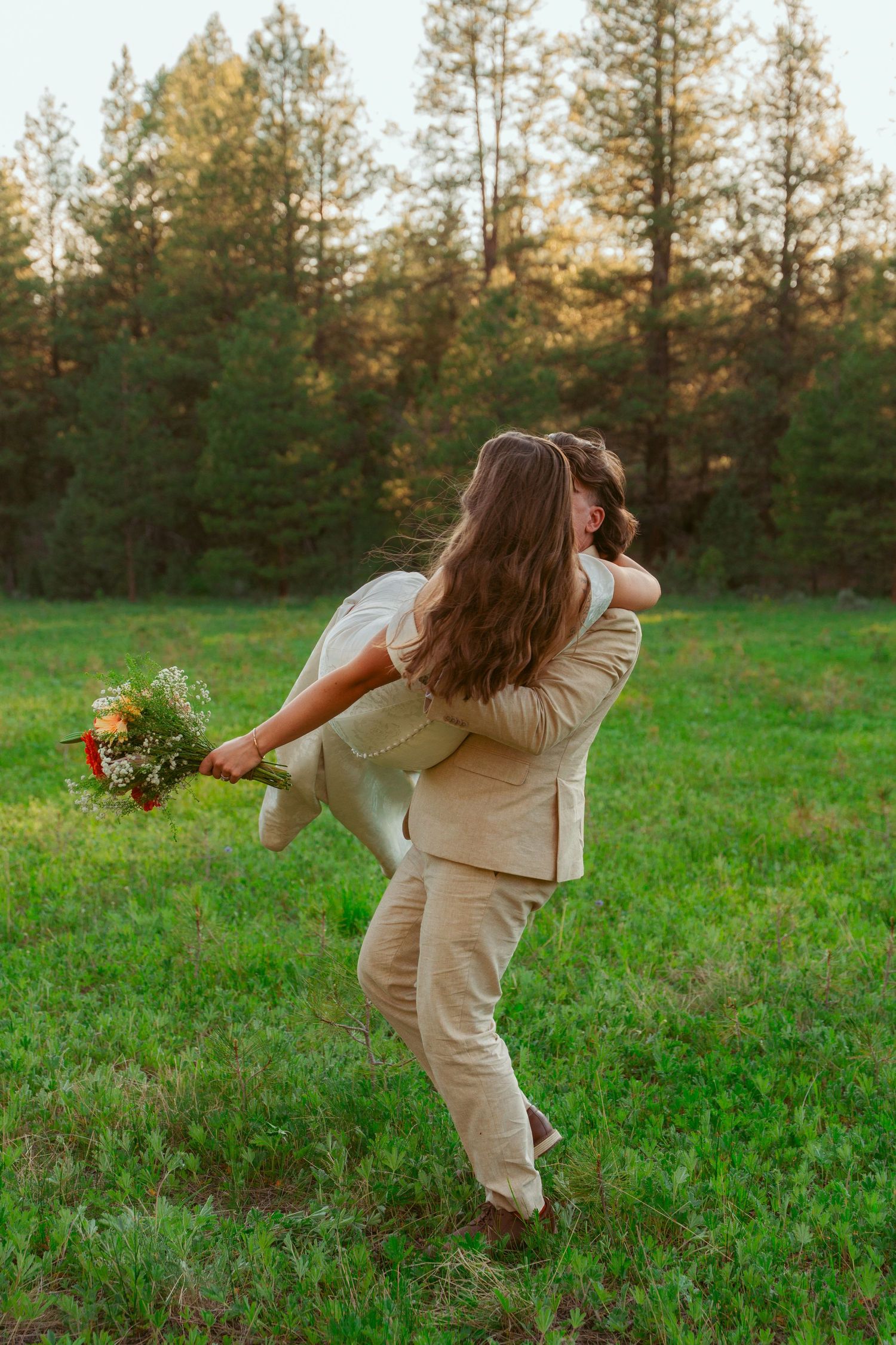 A couple in matching beige outfits dances playfully in a sunlit meadow near a pine forest.