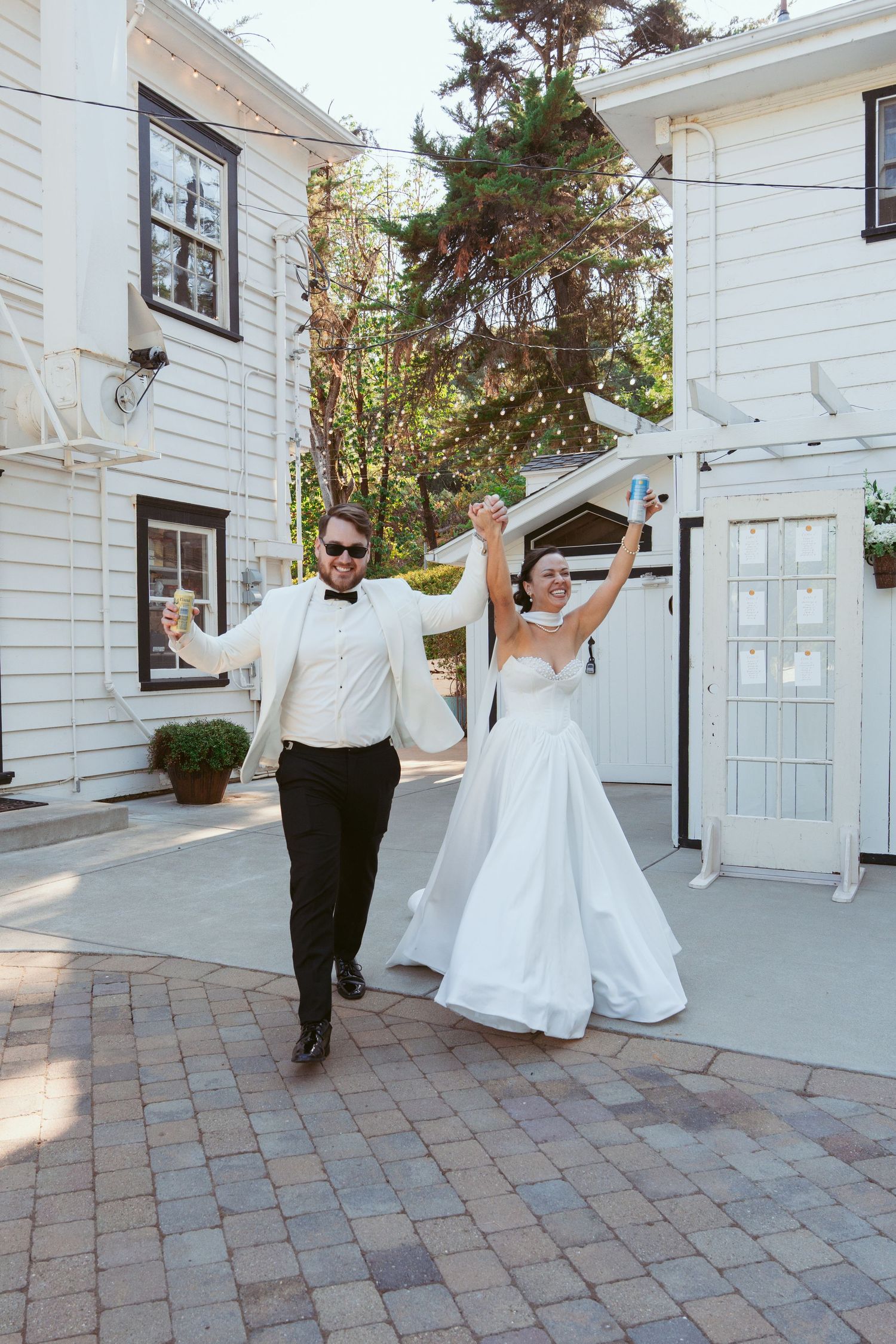 Joyful couple dancing and twirling on brick patio outside white colonial style building.
