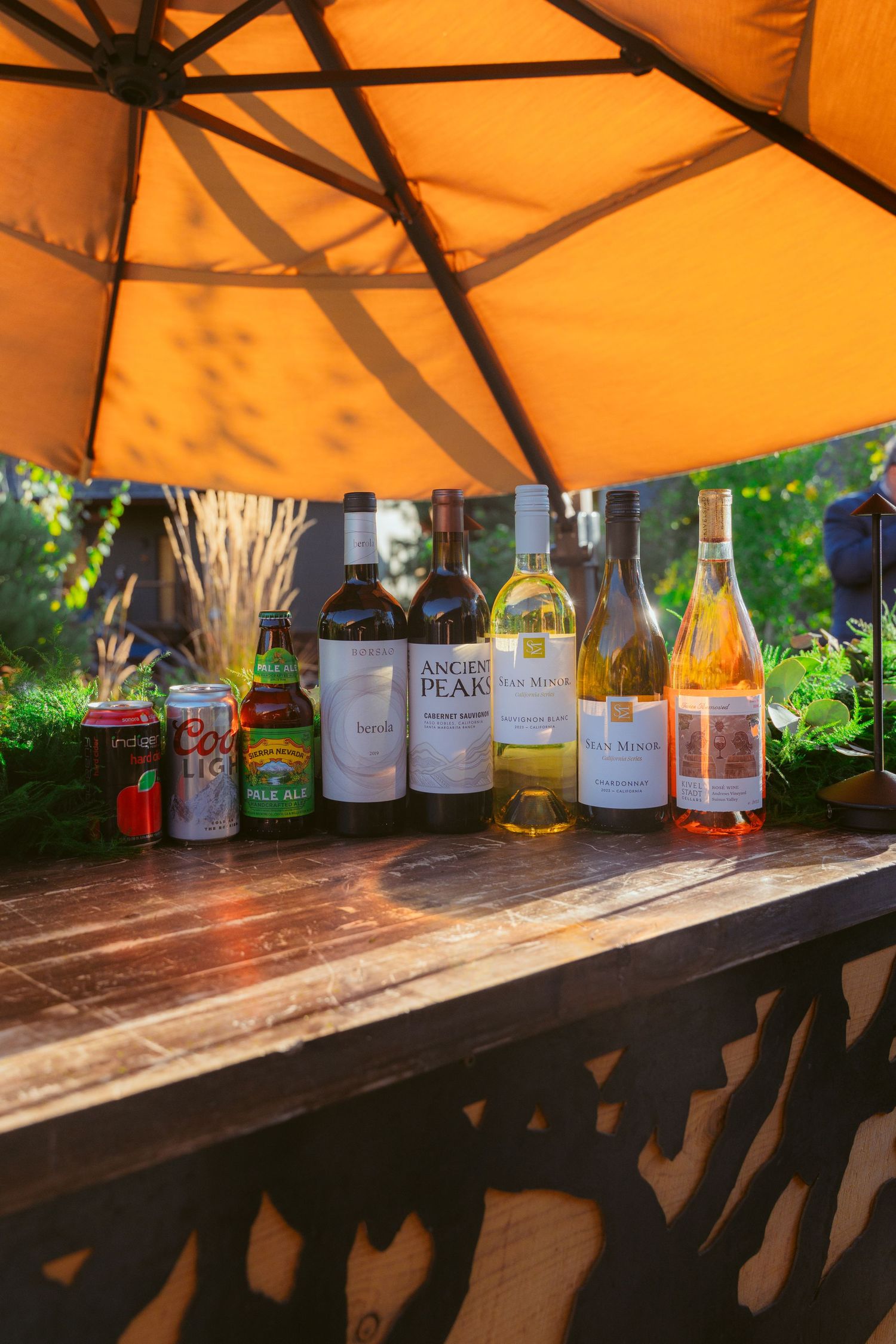 Various wine and liquor bottles displayed on a wooden bar under an orange tent canopy.