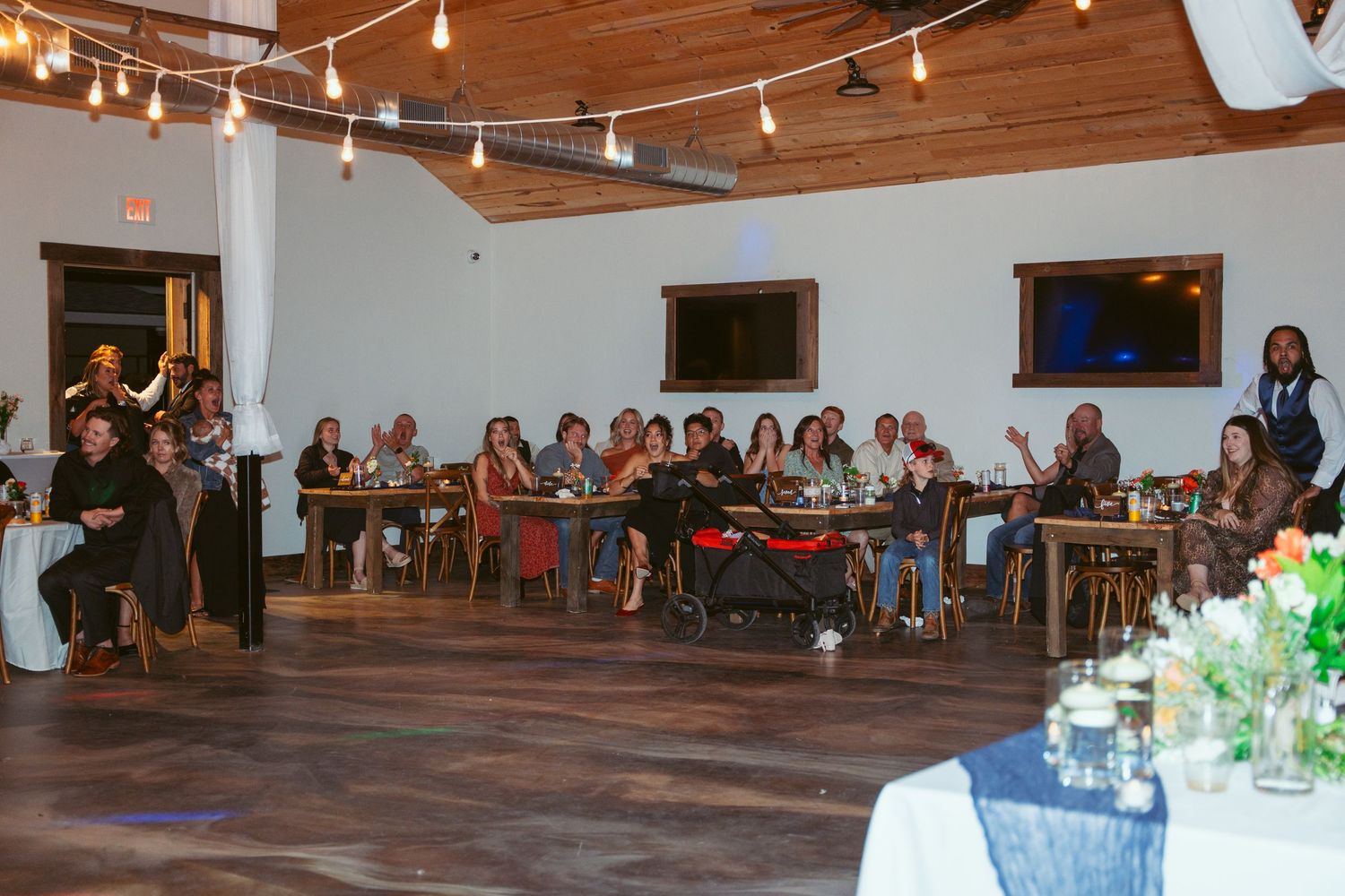 Reception venue filled with guests at wooden tables under string lights and wooden ceiling beams.