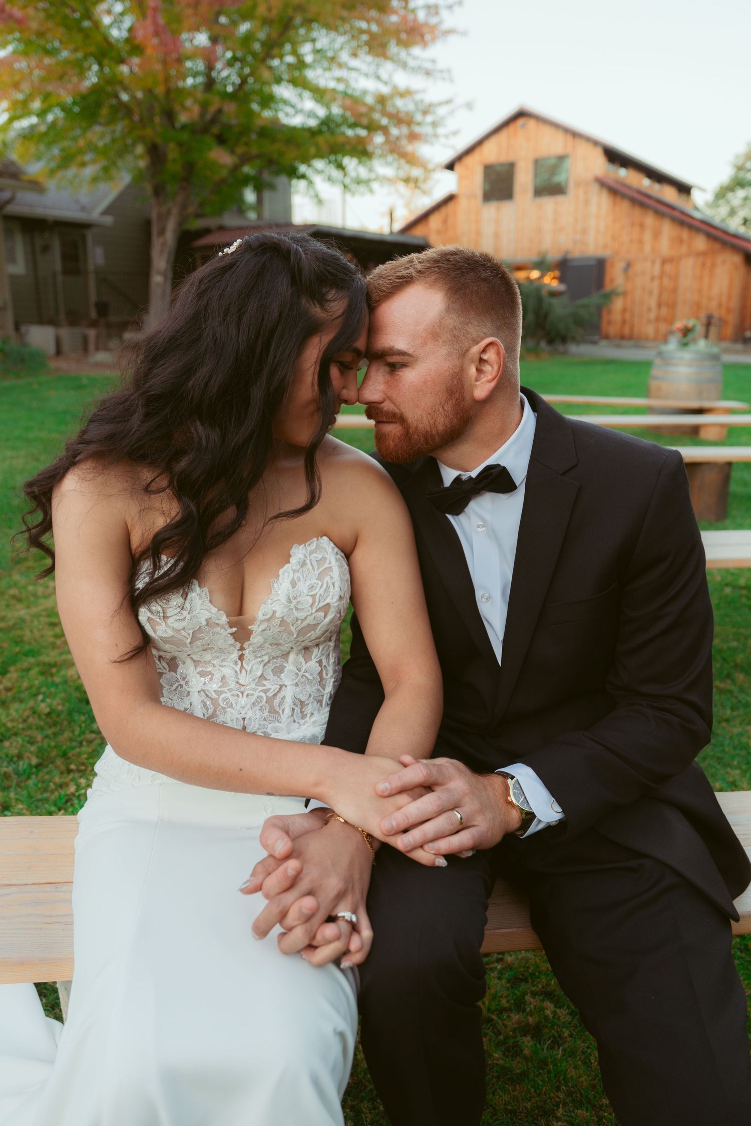 Romantic outdoor wedding couple embraces on rustic wooden deck with barn in background.