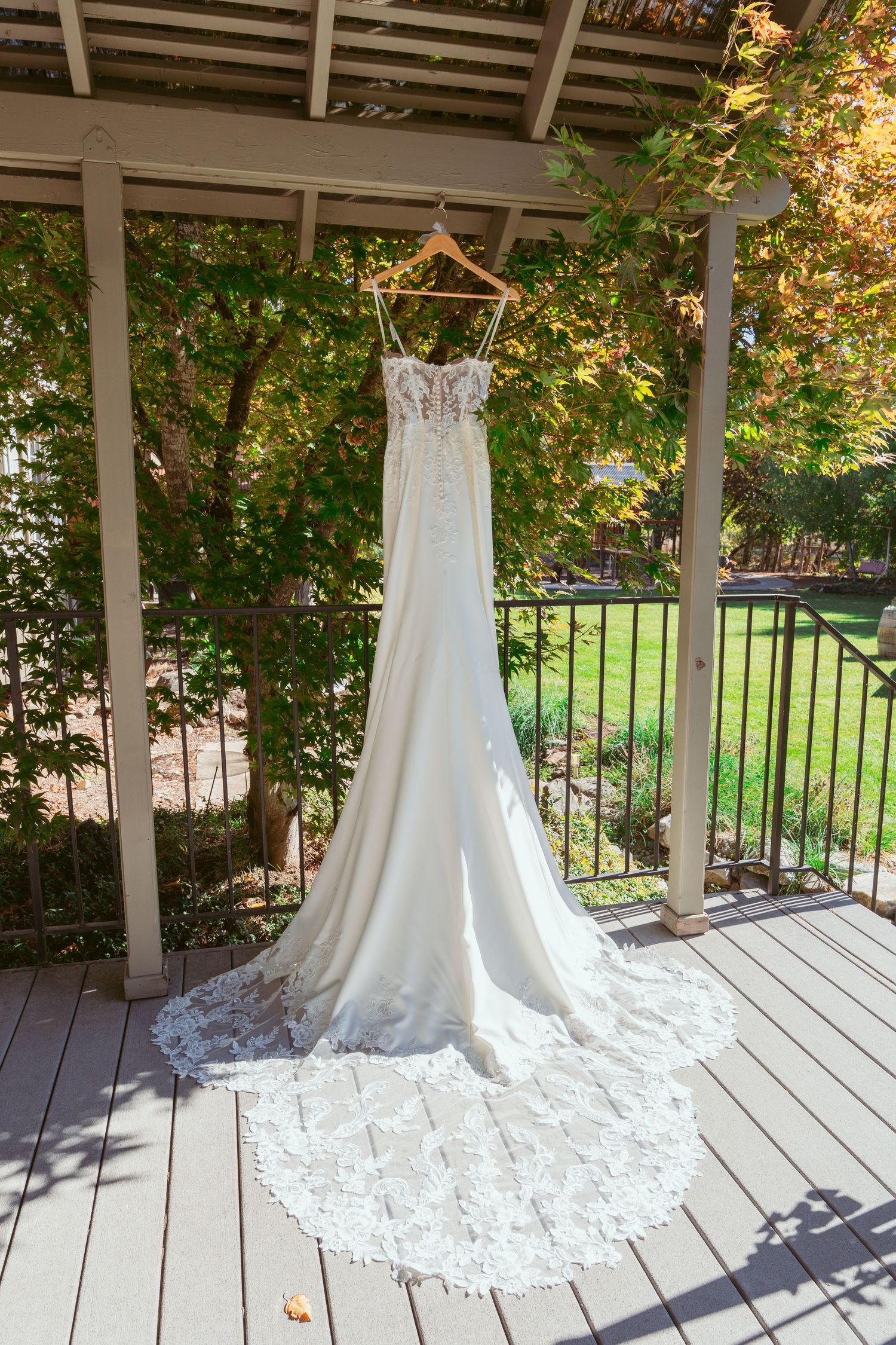 White lace wedding gown with long train displayed on outdoor deck surrounded by greenery.