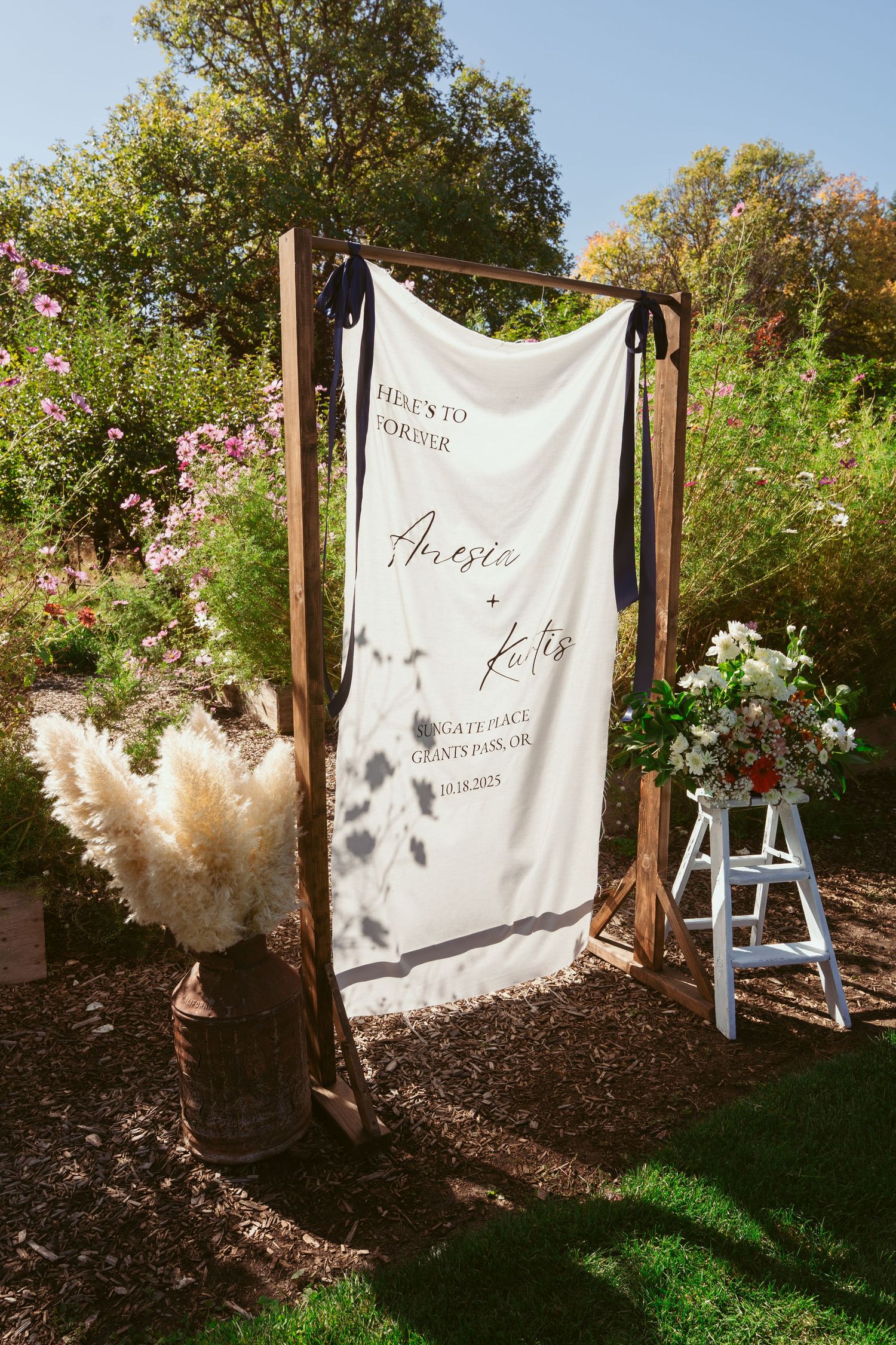 A white backdrop hangs in a garden setting with pampas grass decorations creating a romantic outdoor photo area.
