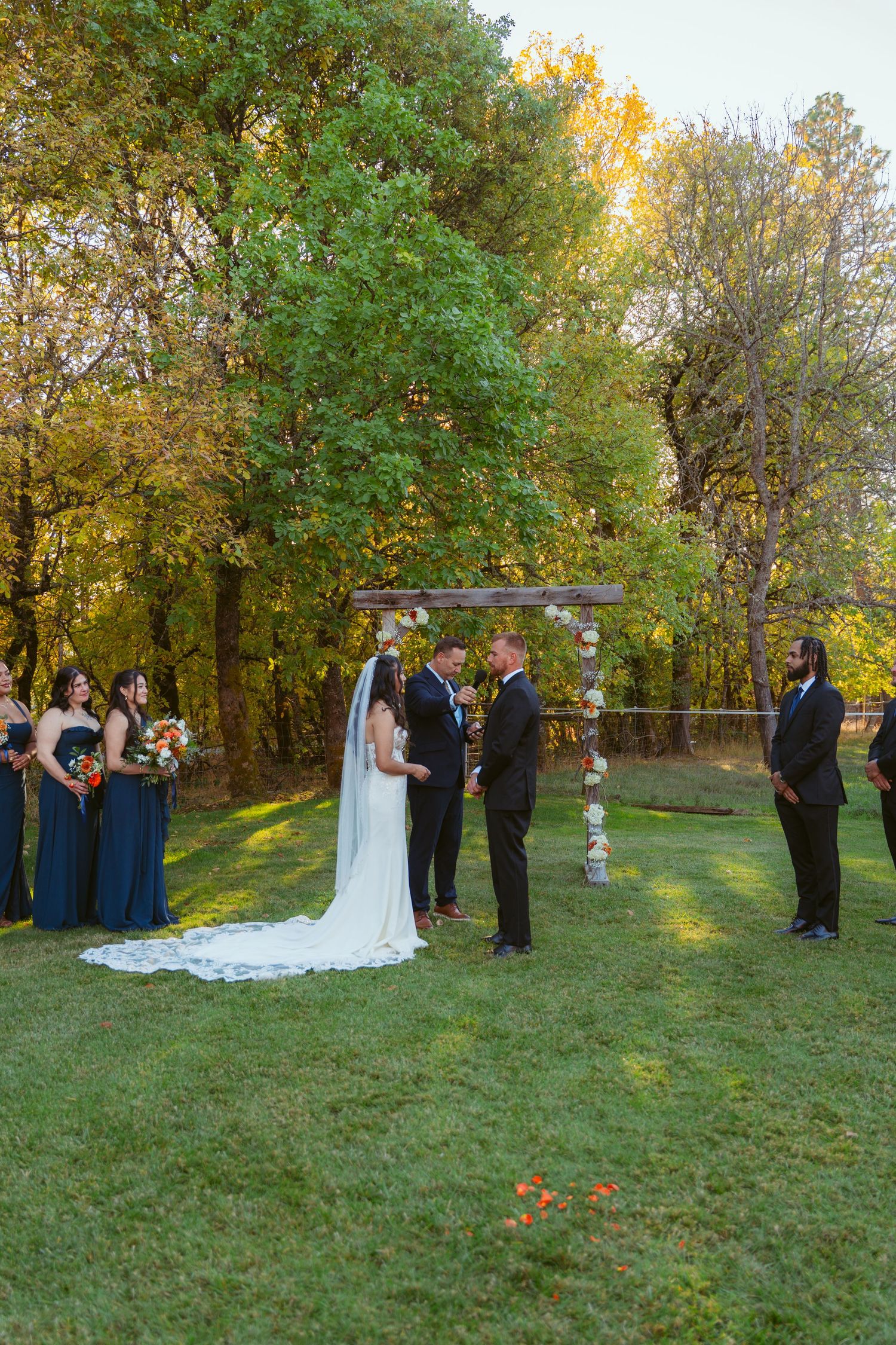 Wedding ceremony setup with floral arch and bridal party against autumn trees.