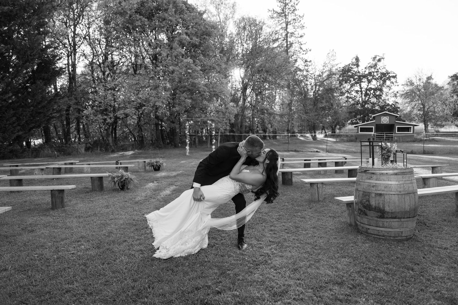 Black and white photo of wedding couple in dramatic dip pose among rustic outdoor seating.