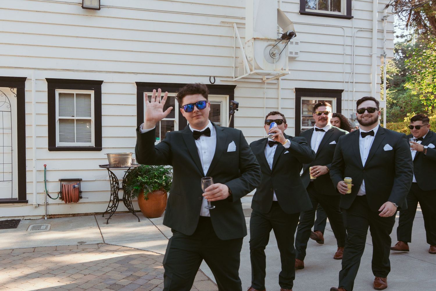 Groomsmen in black tuxedos walking and posing together outside a white building.