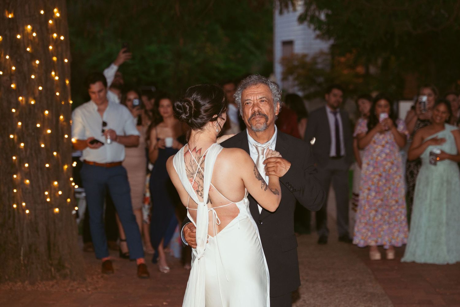 Couples dance at dusk under string lights at an outdoor wedding reception with guests watching.