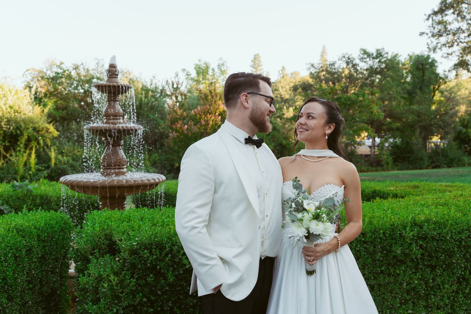 Bride and groom share an intimate moment in a garden setting with a decorative fountain in the background.