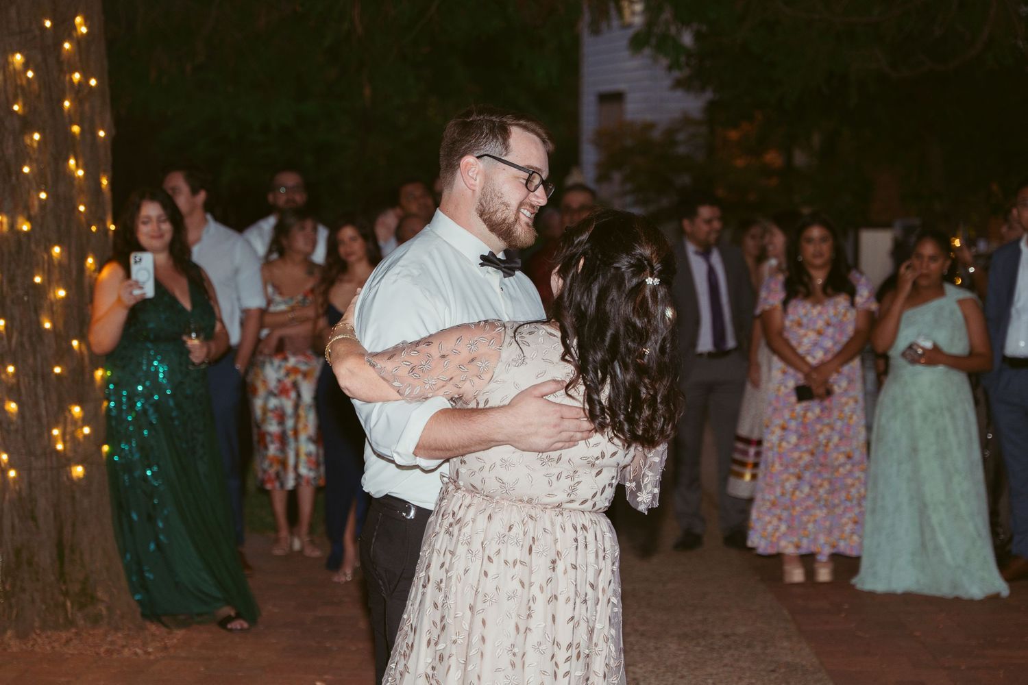 Couple dances surrounded by guests in formal attire at outdoor evening reception.