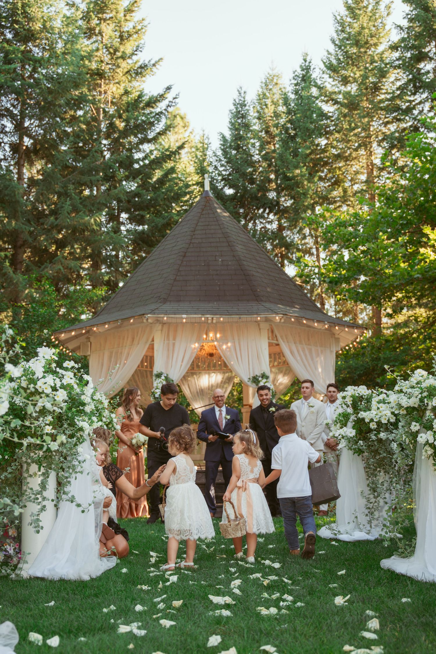 Wedding guests gather for an outdoor ceremony under tall pine trees with white floral decorations and a gazebo.