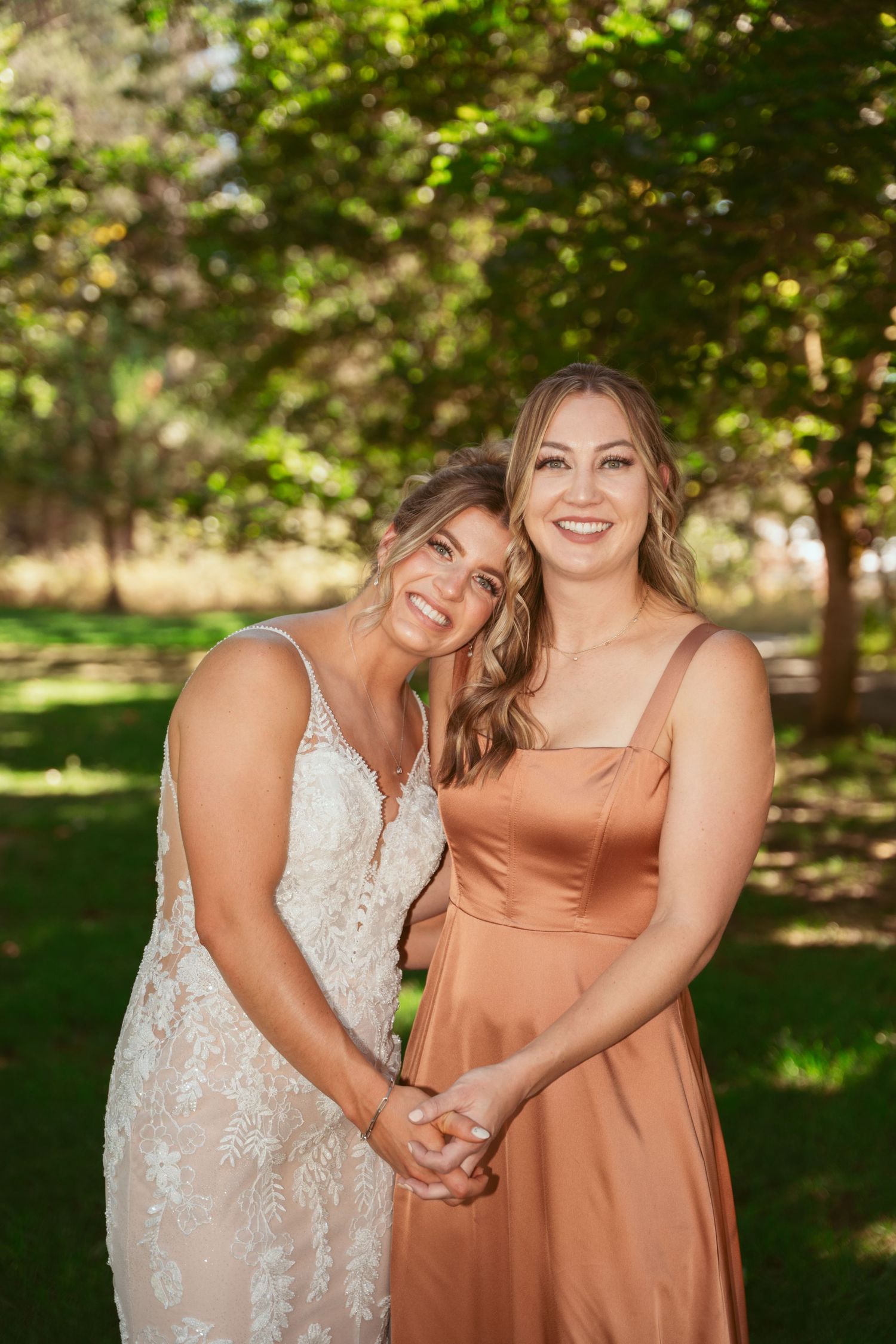 Two people in complementary formal dresses embrace while laughing together in a sunlit outdoor setting.