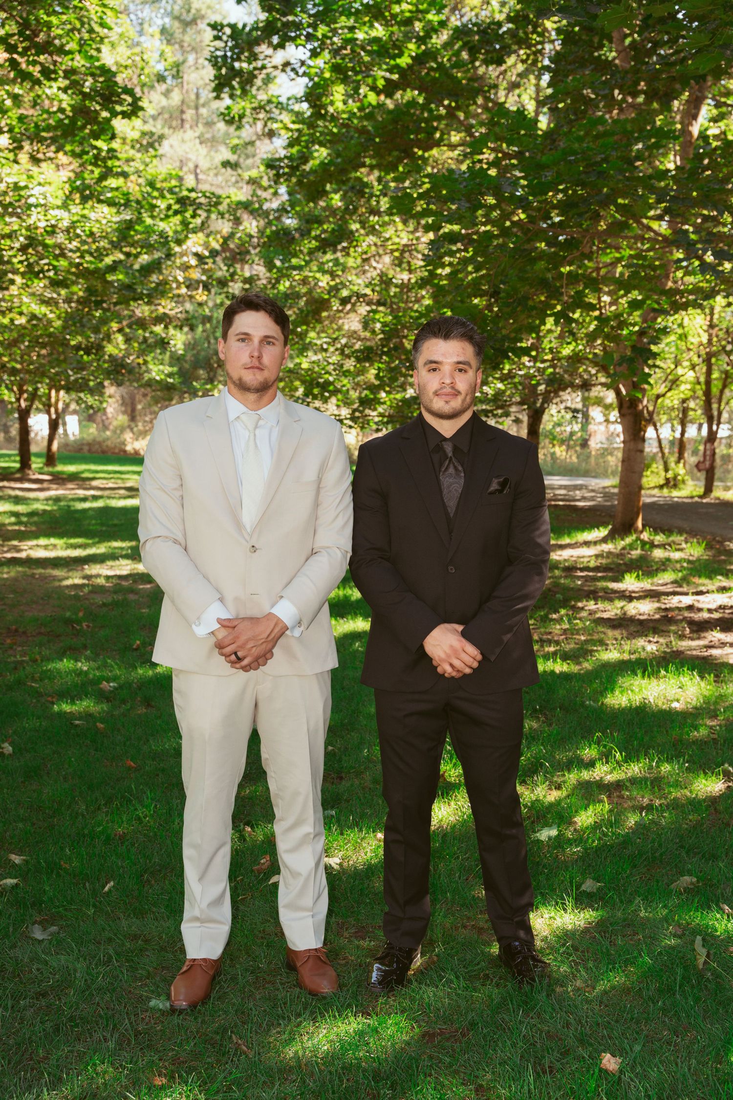 Two groomsmen in white and black suits stand together on a sunny lawn.