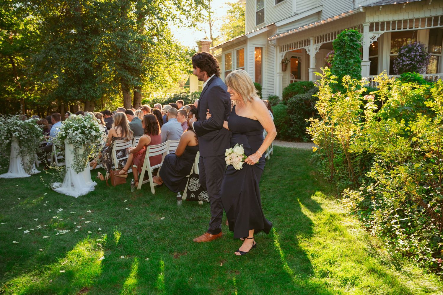 Guests gather on a lawn outside a Victorian-style house during a sunny outdoor event.
