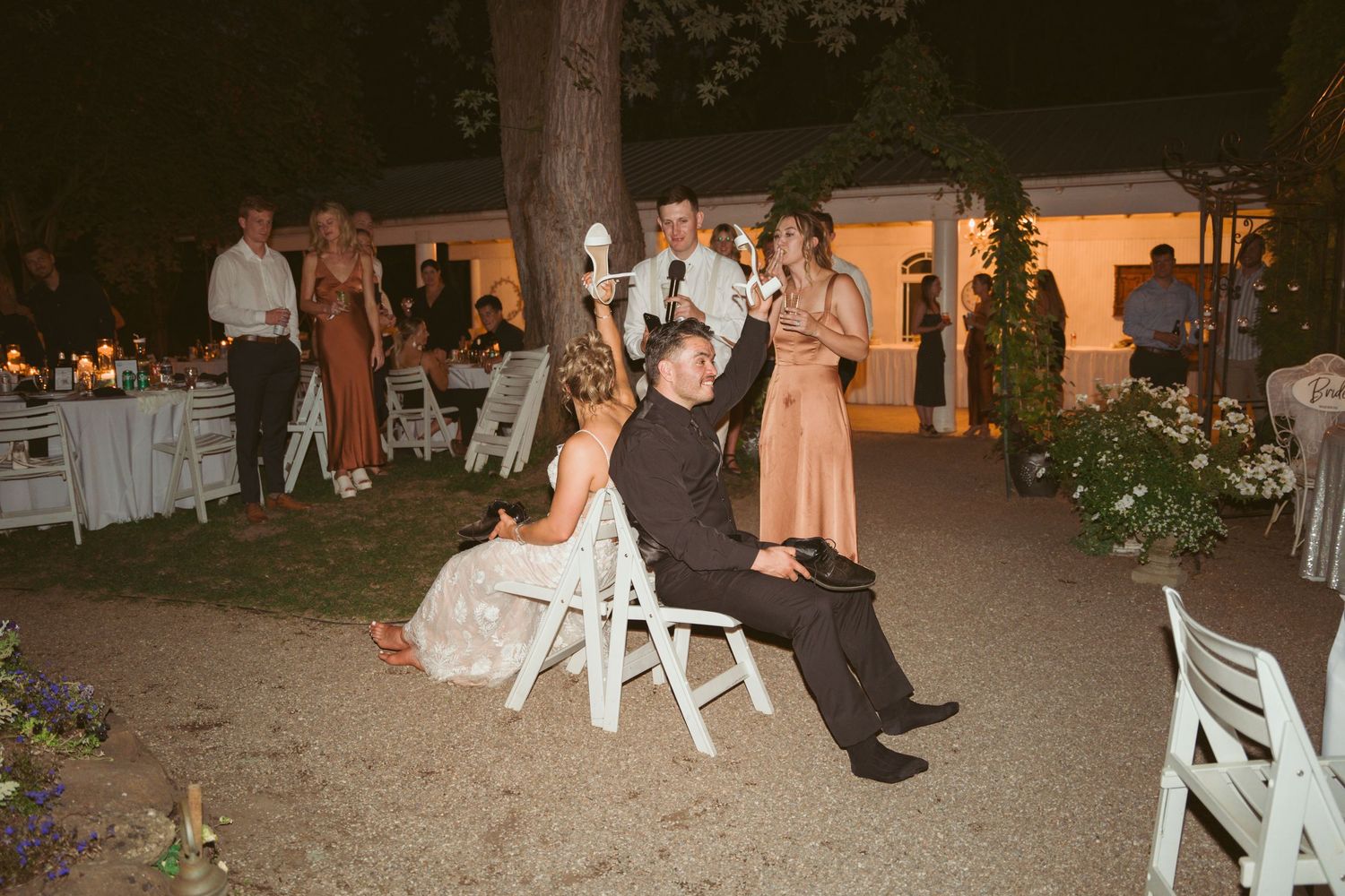 Guests celebrating at outdoor wedding reception with white chairs and string lighting visible.