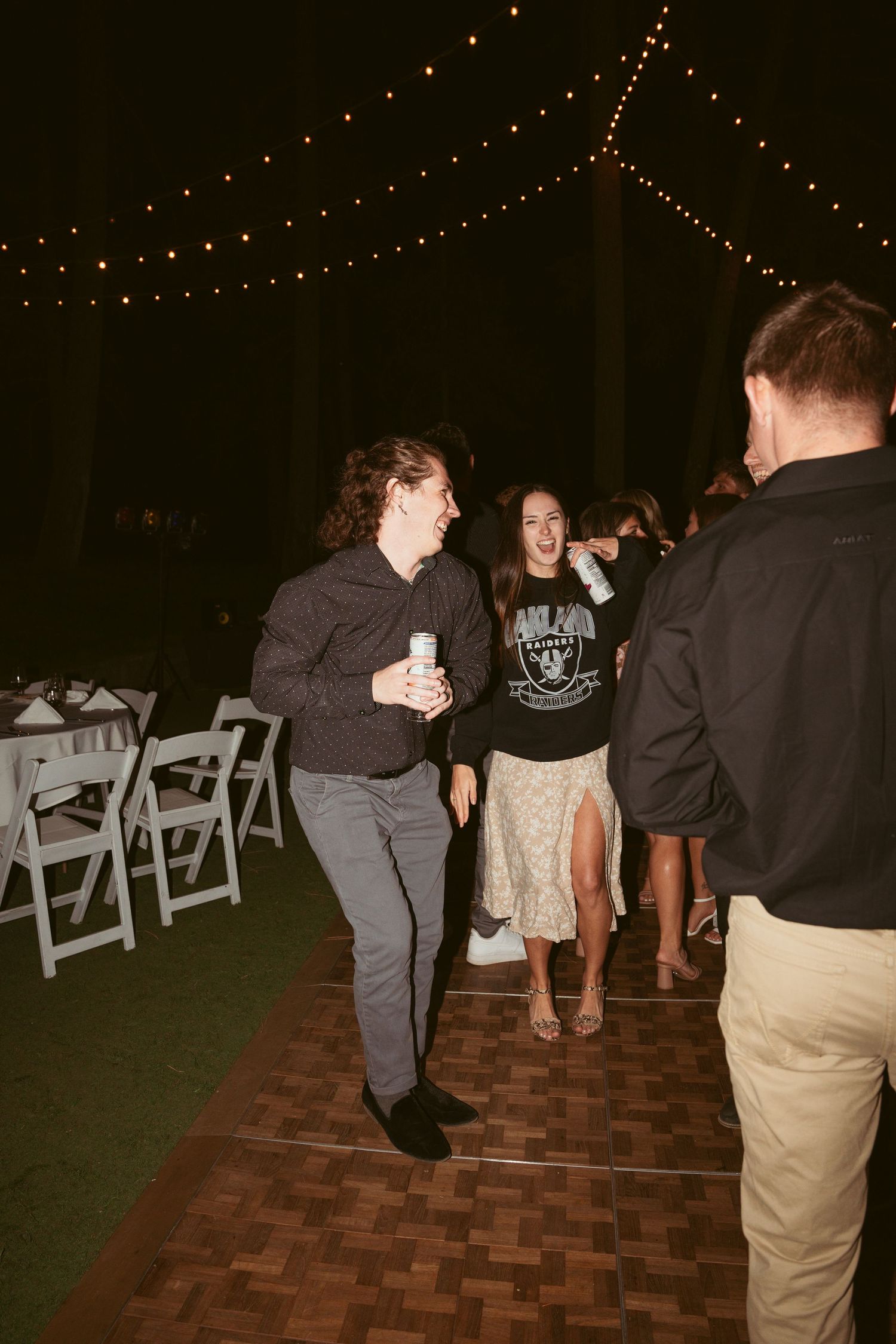 People dancing on a brick patio at night with folding chairs and string lights visible.