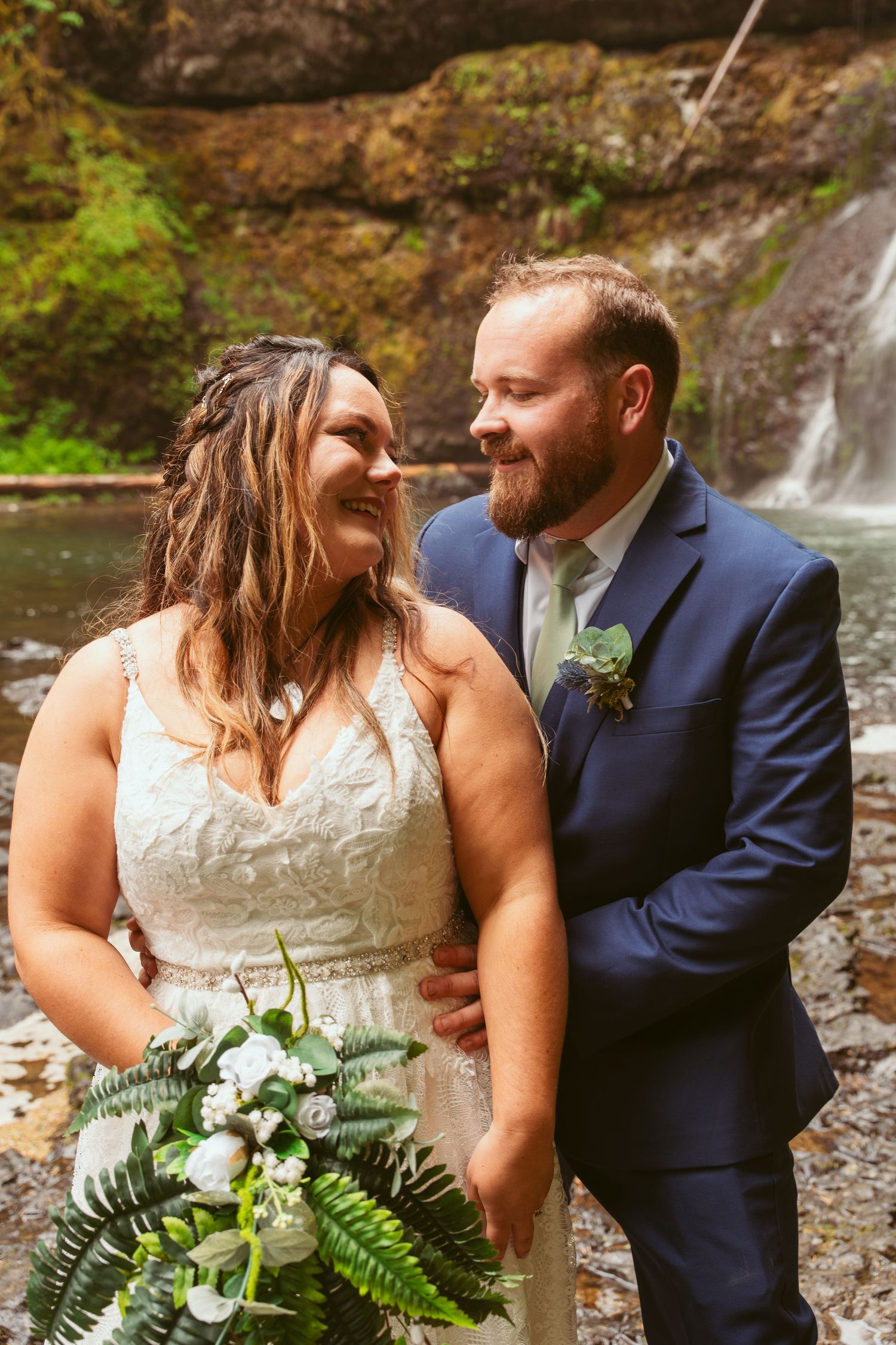 Newlyweds share an intimate moment by a waterfall, bride holding a fern bouquet.