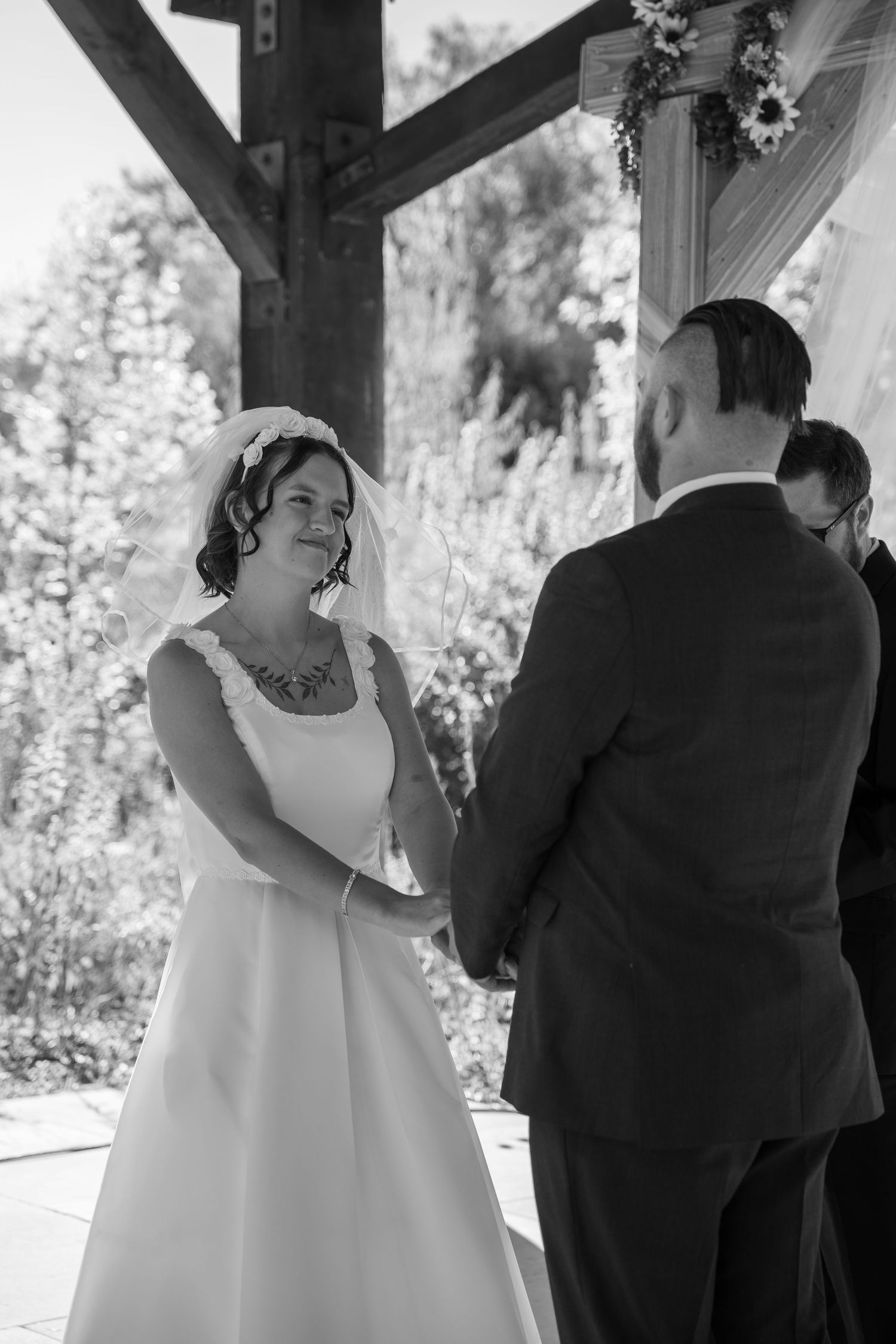 A black and white wedding ceremony moment captured under a rustic wooden pergola with floral decorations.