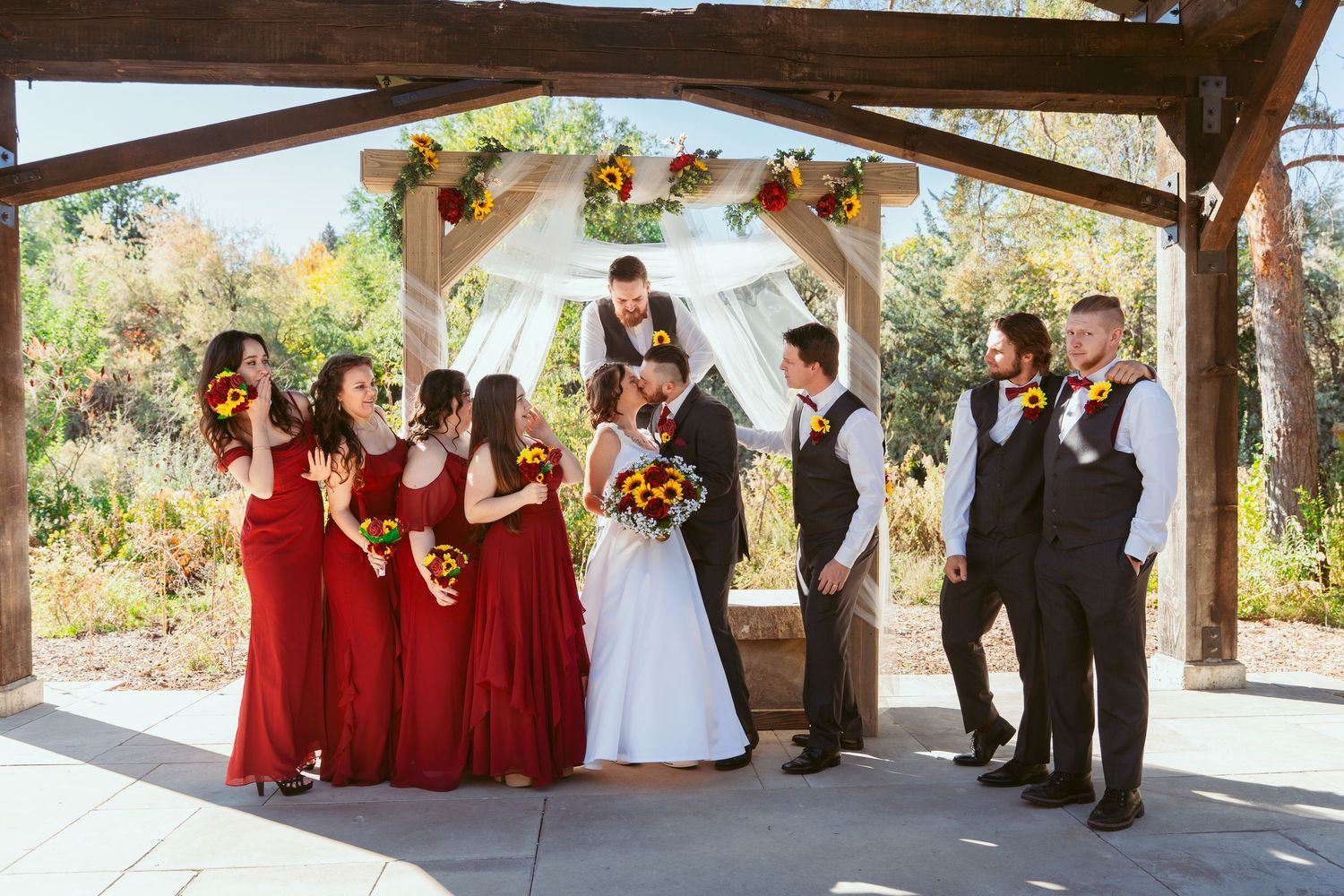 Full wedding party group photo showing bridesmaids in red and groomsmen in black under pavilion.
