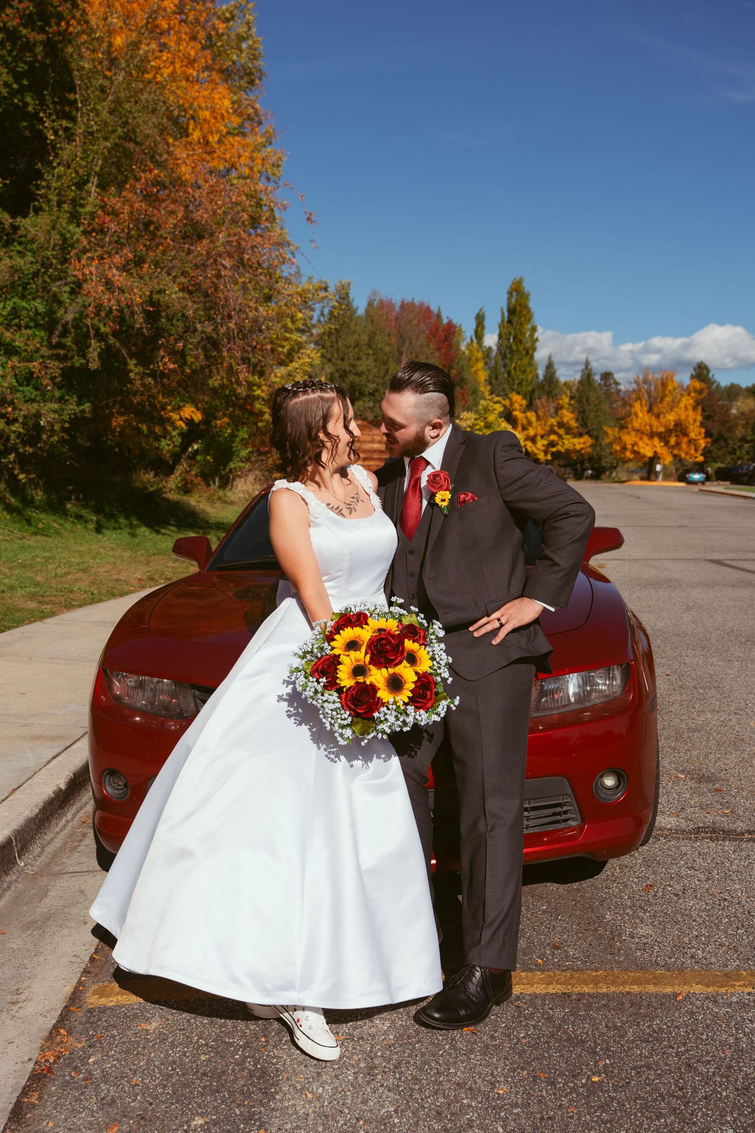 Autumn wedding photoshoot with couple and red car against colorful fall foliage.