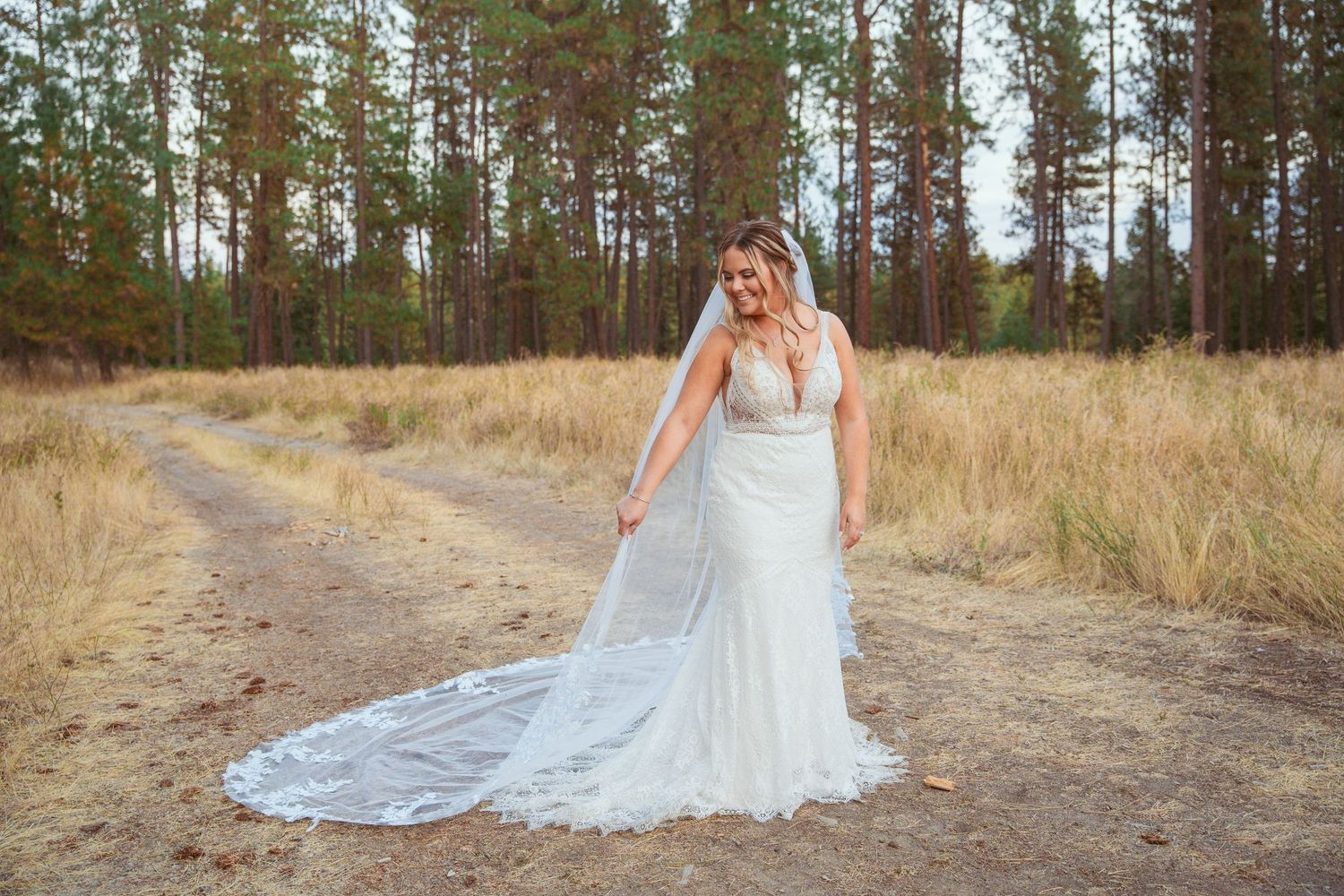 Lace wedding dress with long train photographed on dirt path in pine forest.