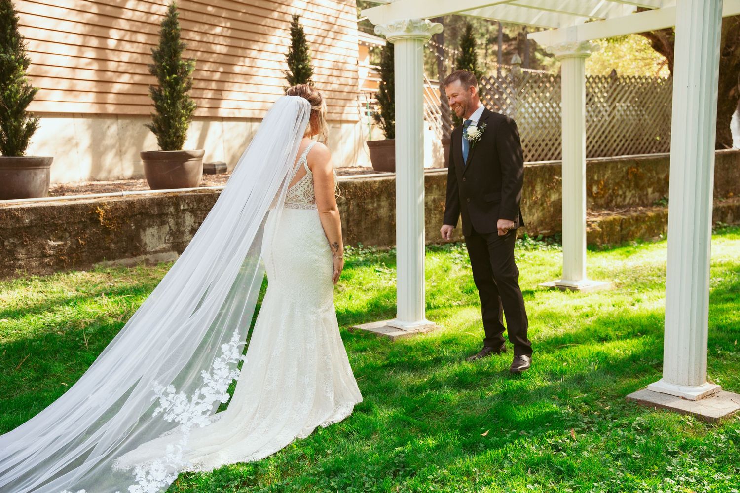 First look moment between a bride in a long veil and groom by white garden pillars on a sunny day.