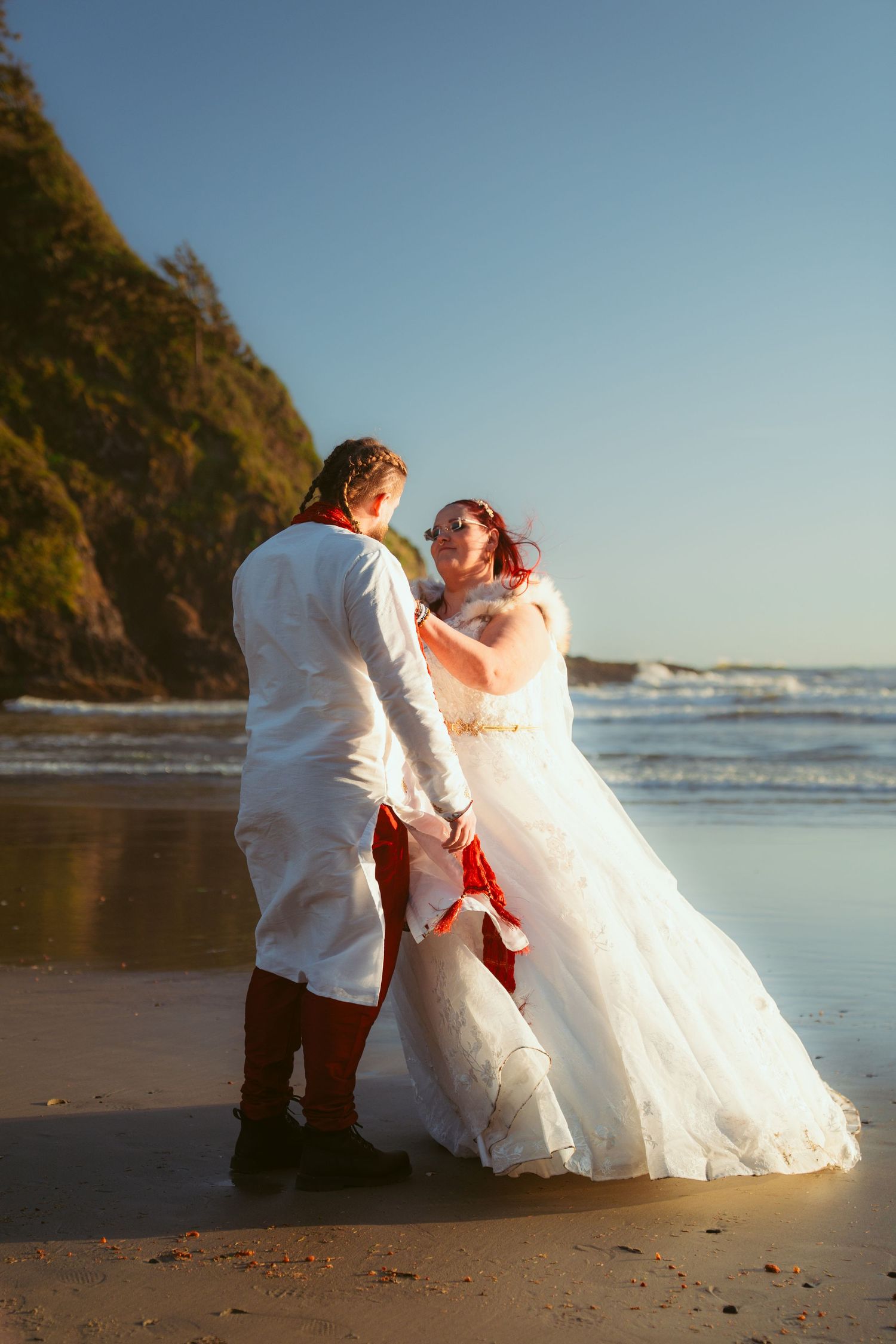 A couple in wedding attire dancing on a beach at sunset with cliffs in the background.