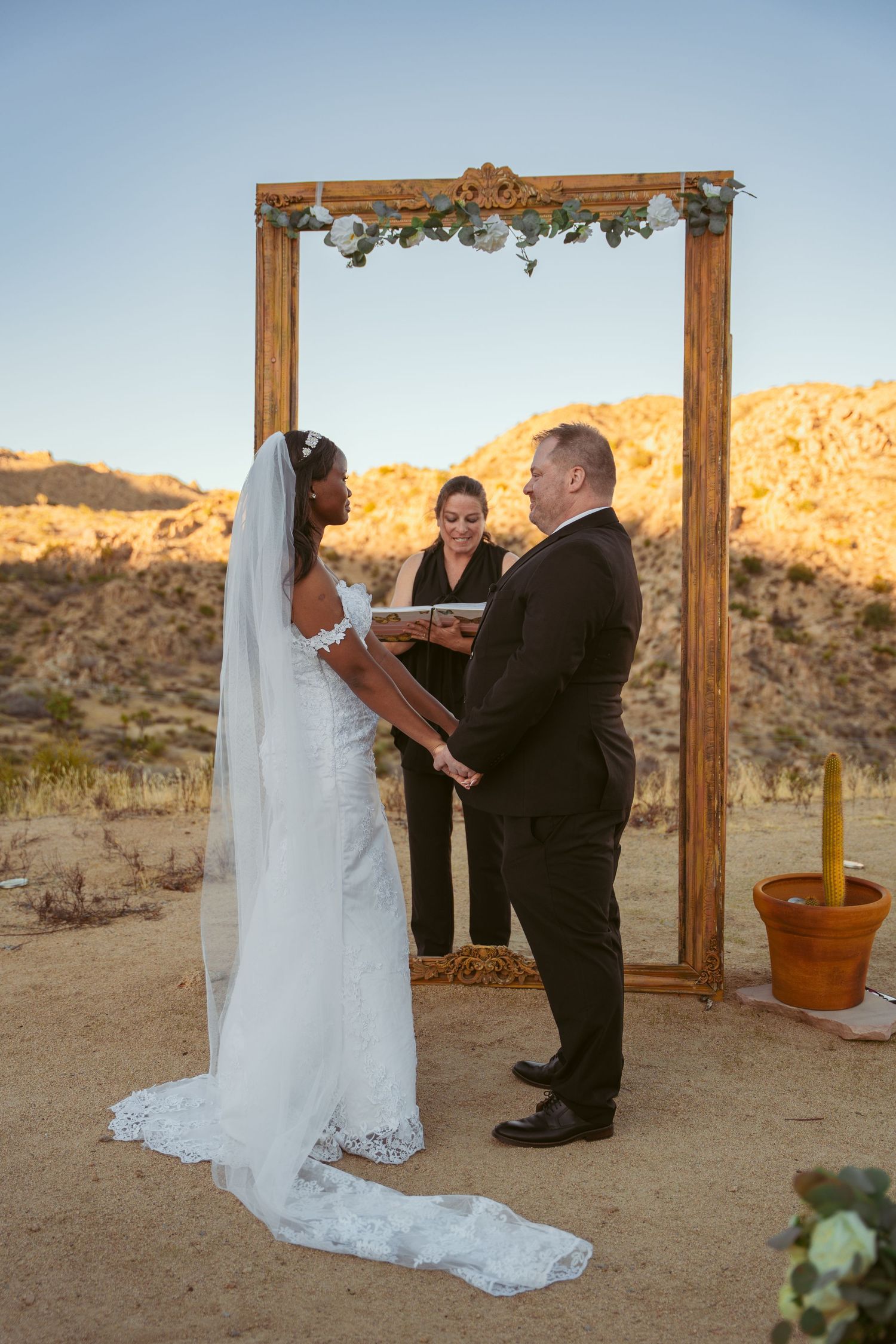 Couple exchanging vows during desert wedding ceremony beneath ornate wooden arch frame.